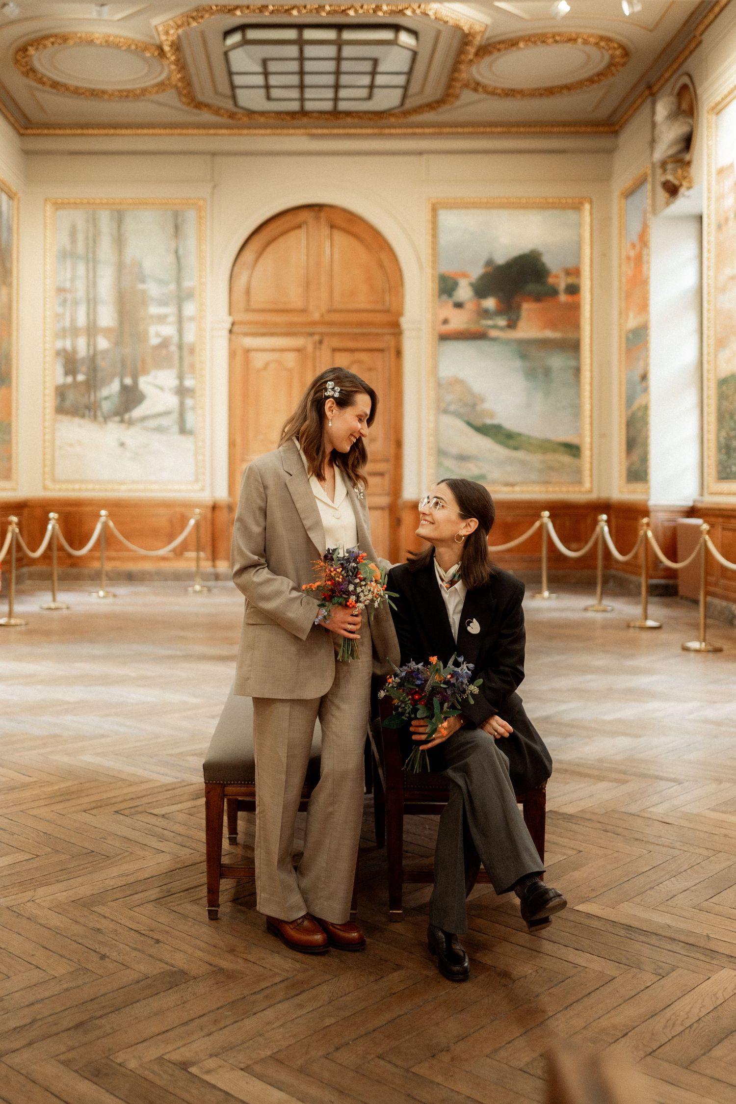 Two women in elegant suits hold bouquets and share a tender moment in a grand ornate European hall.