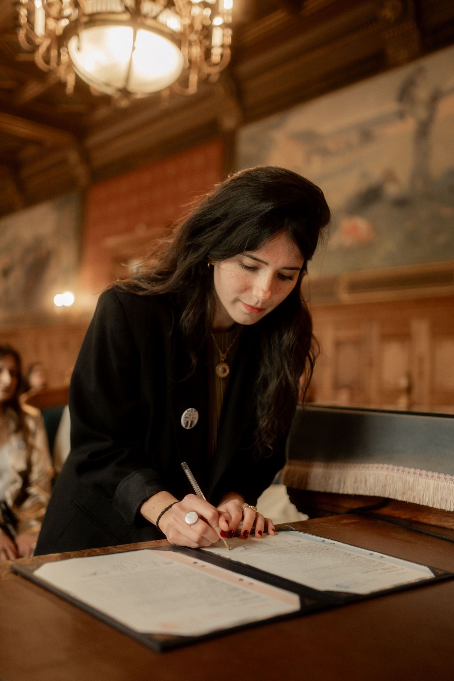 Bride in black blazer signs wedding certificate at ornate wooden table in chandelier-lit room.