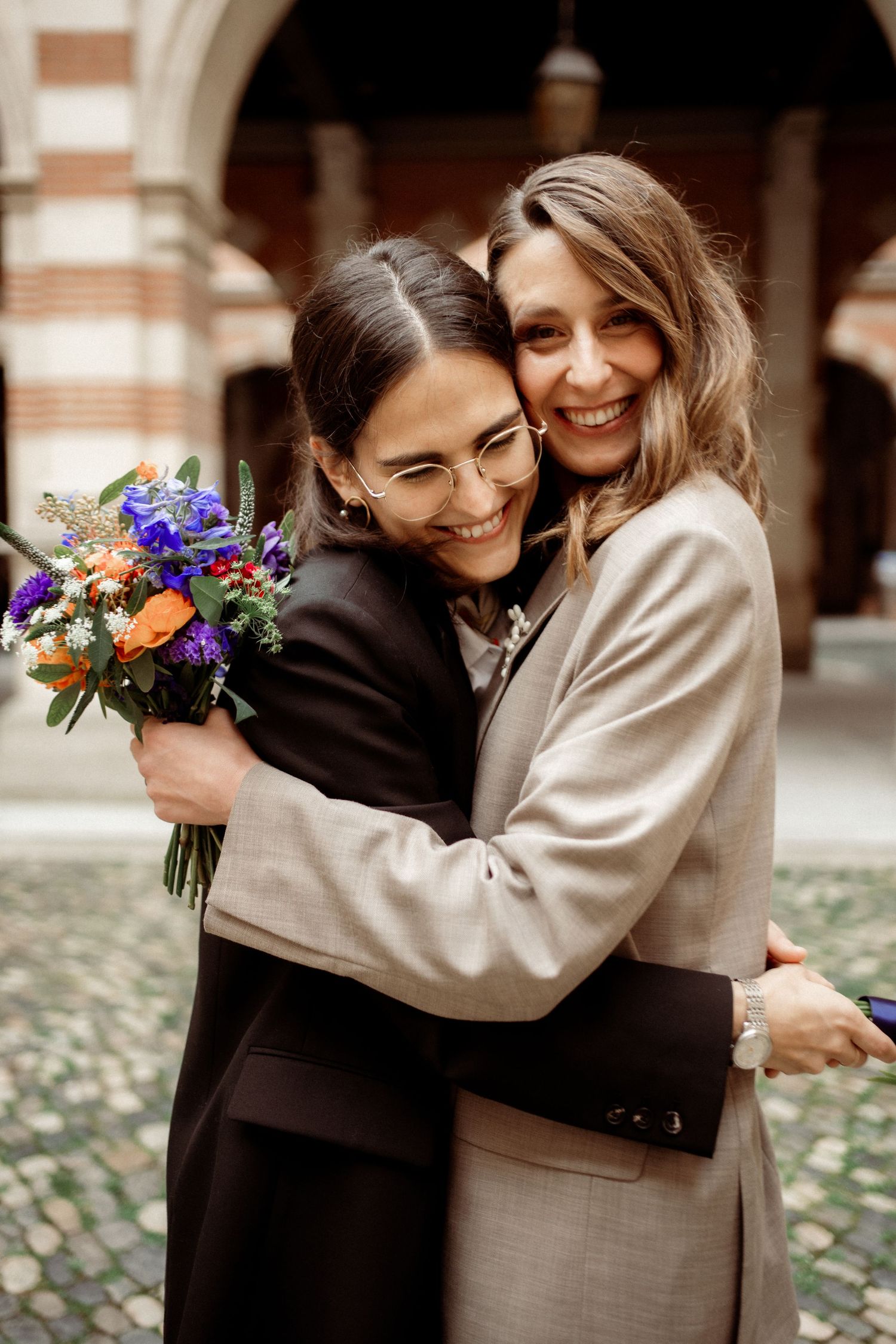 Two women hugging and smiling, one holding a colorful bouquet, in an elegant courtyard setting.