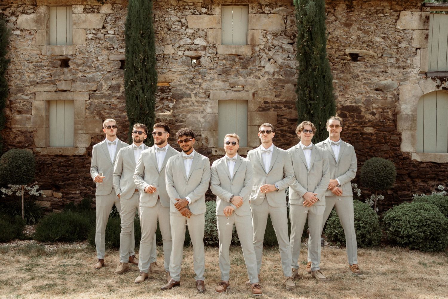 A wedding party in light colored suits poses against a rustic stone wall with cypress trees in the background.