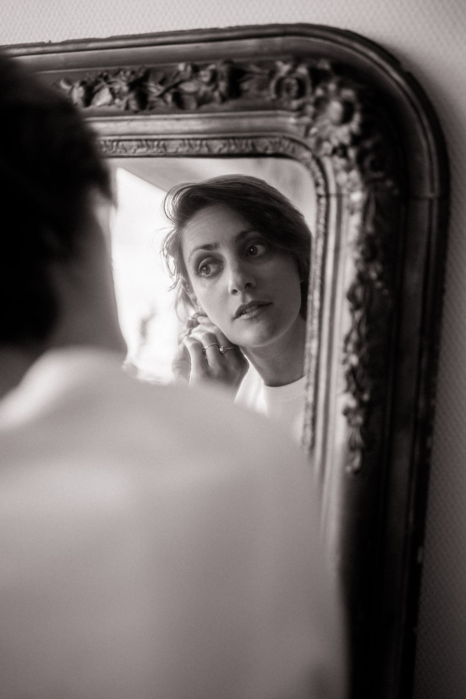 Woman applying earring while gazing intently at her reflection in an ornate vintage mirror, black and white photo.