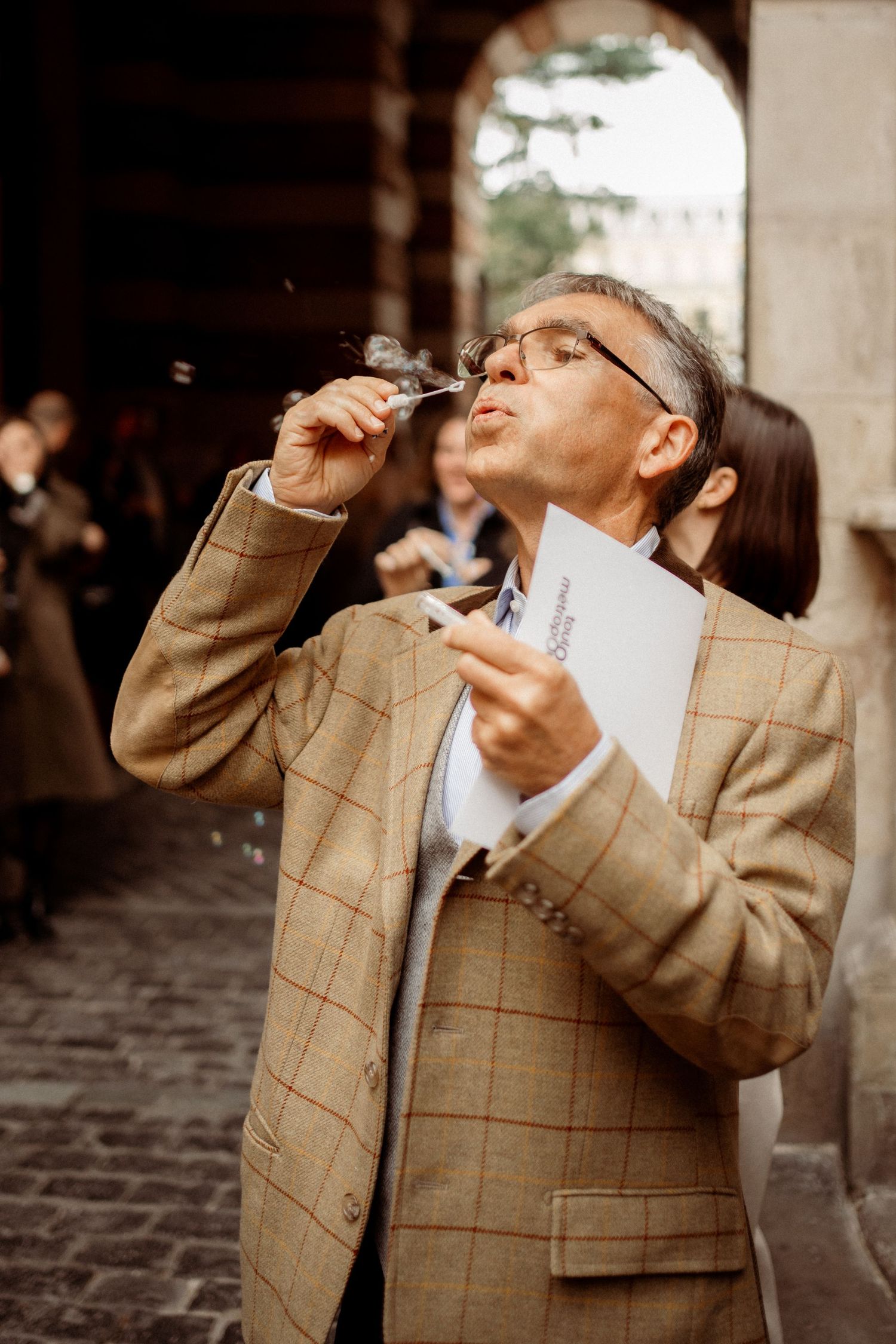 Older man in plaid blazer blowing soap bubbles outdoors near historic stone archway.