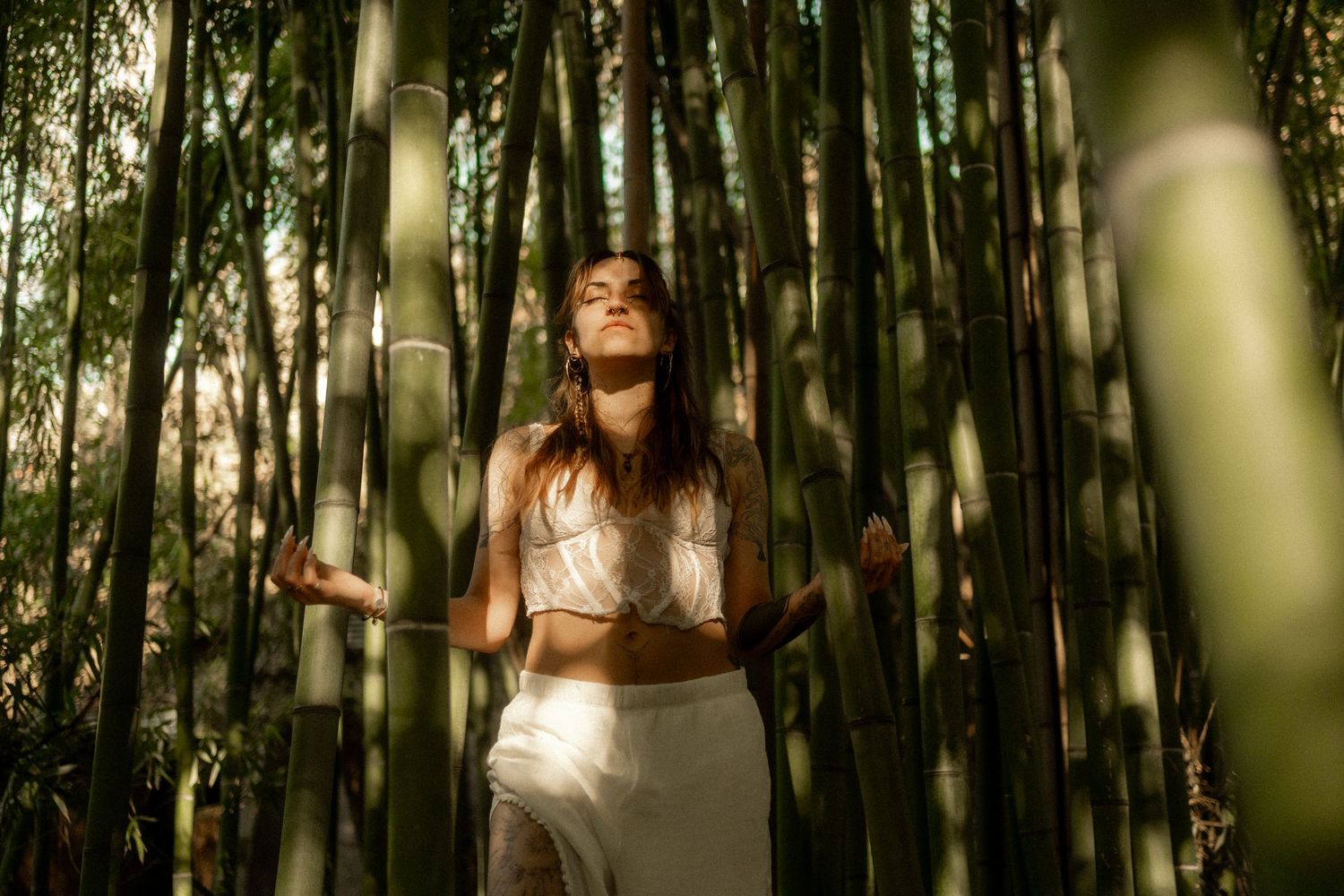 Woman meditating in a lush bamboo forest, bathed in warm sunlight, wearing a white crop top and skirt.