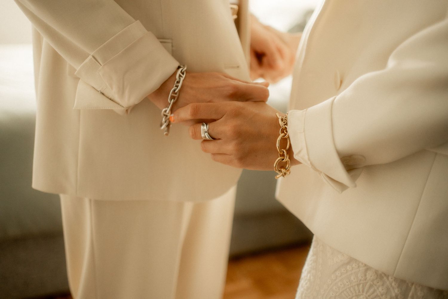 Two women in cream suits clasp hands, wearing gold chain bracelets and a diamond ring, in an elegant bridal preparation moment.