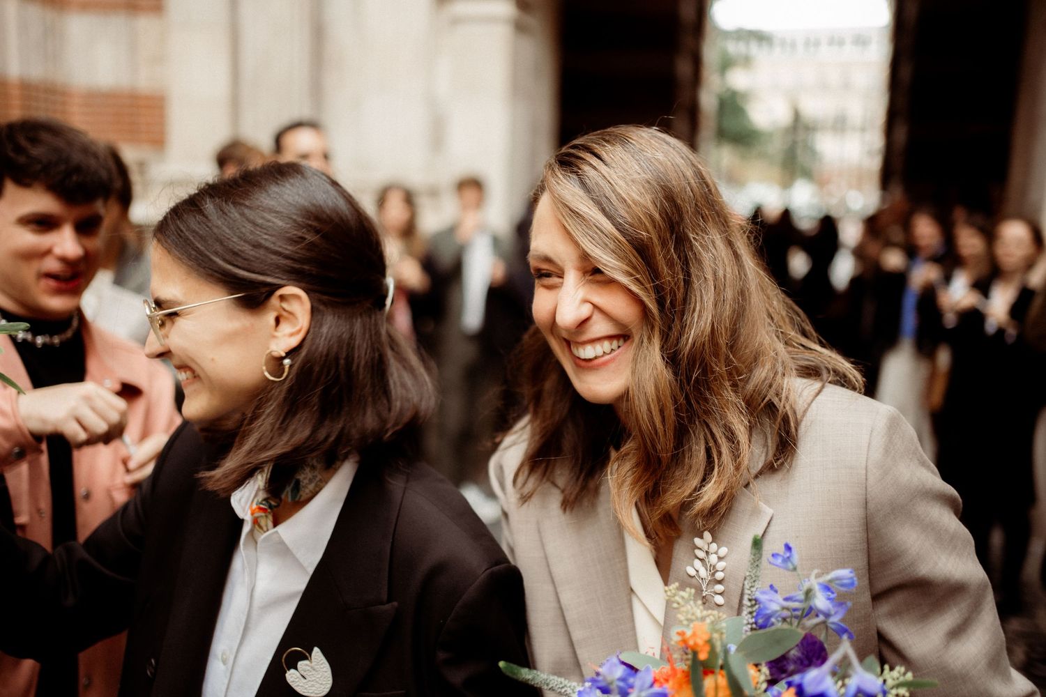 Two smiling women in suits laugh together outdoors, one holding a vibrant bouquet of colorful flowers.