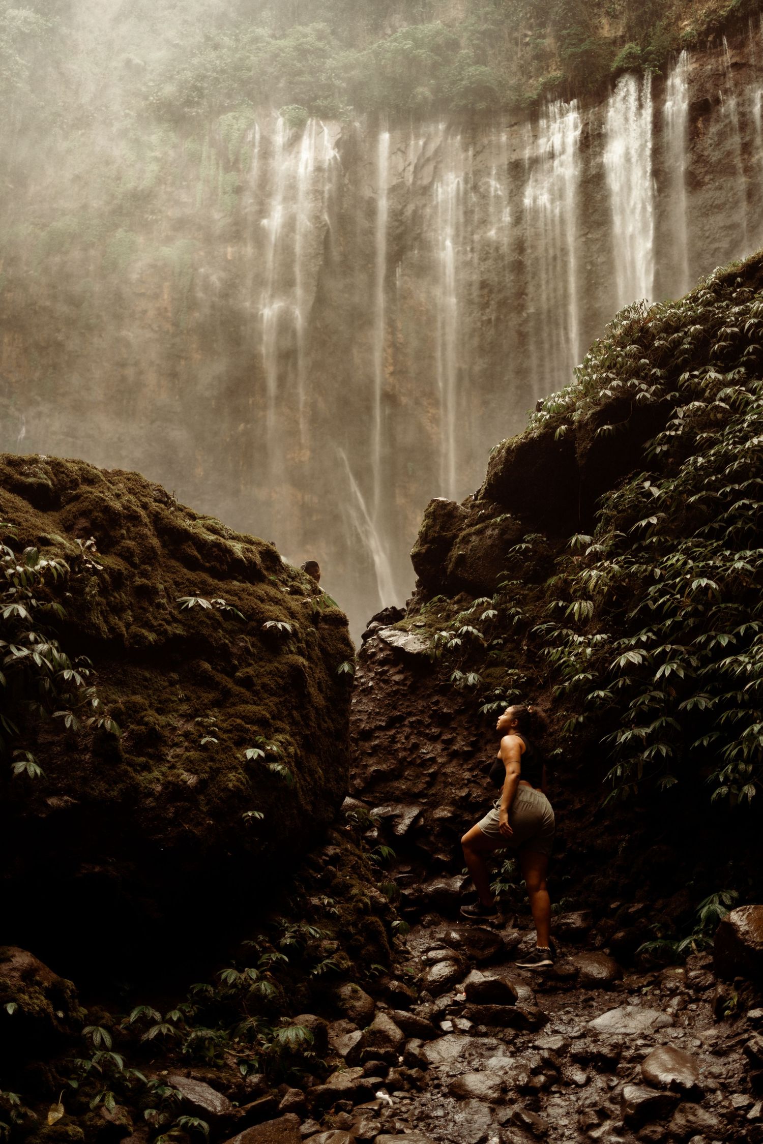 Solo hiker standing before a dramatic misty waterfall cascading down rocky cliffs surrounded by lush vegetation.
