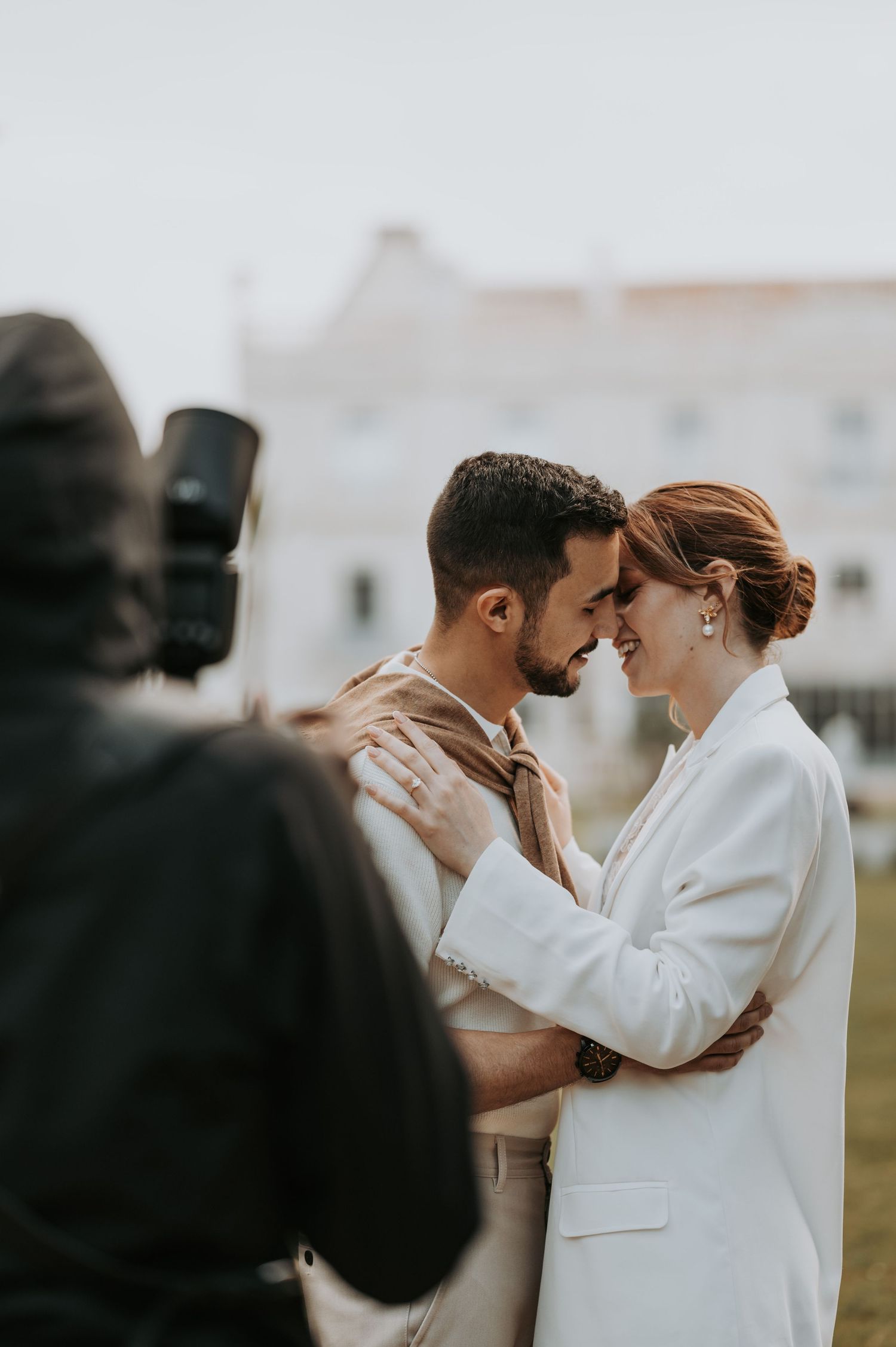 A romantic couple embraces while being photographed, with the photographer visible in silhouette in the foreground.