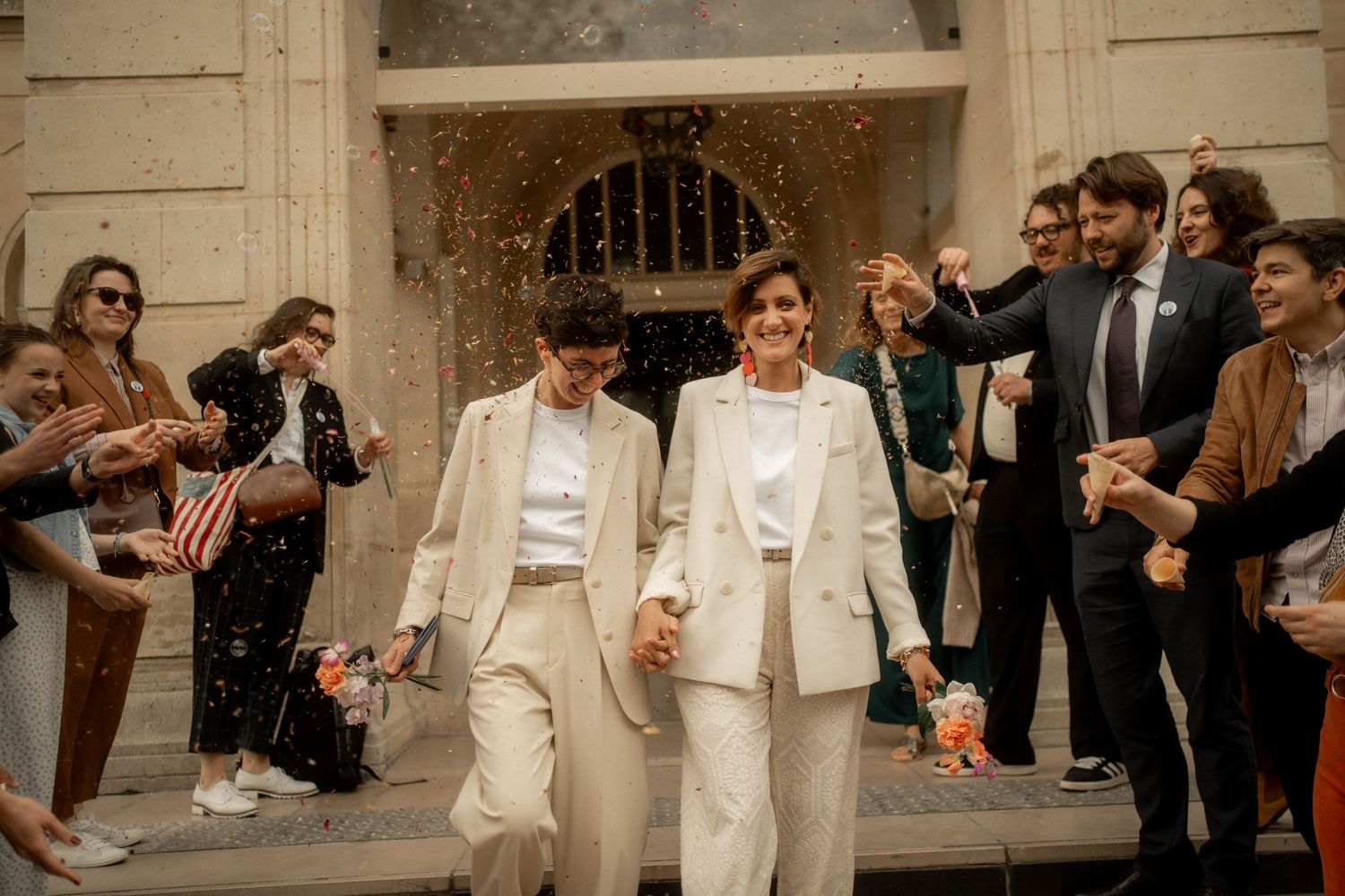 Two brides in white suits walk hand-in-hand as guests celebrate with confetti at their wedding ceremony.