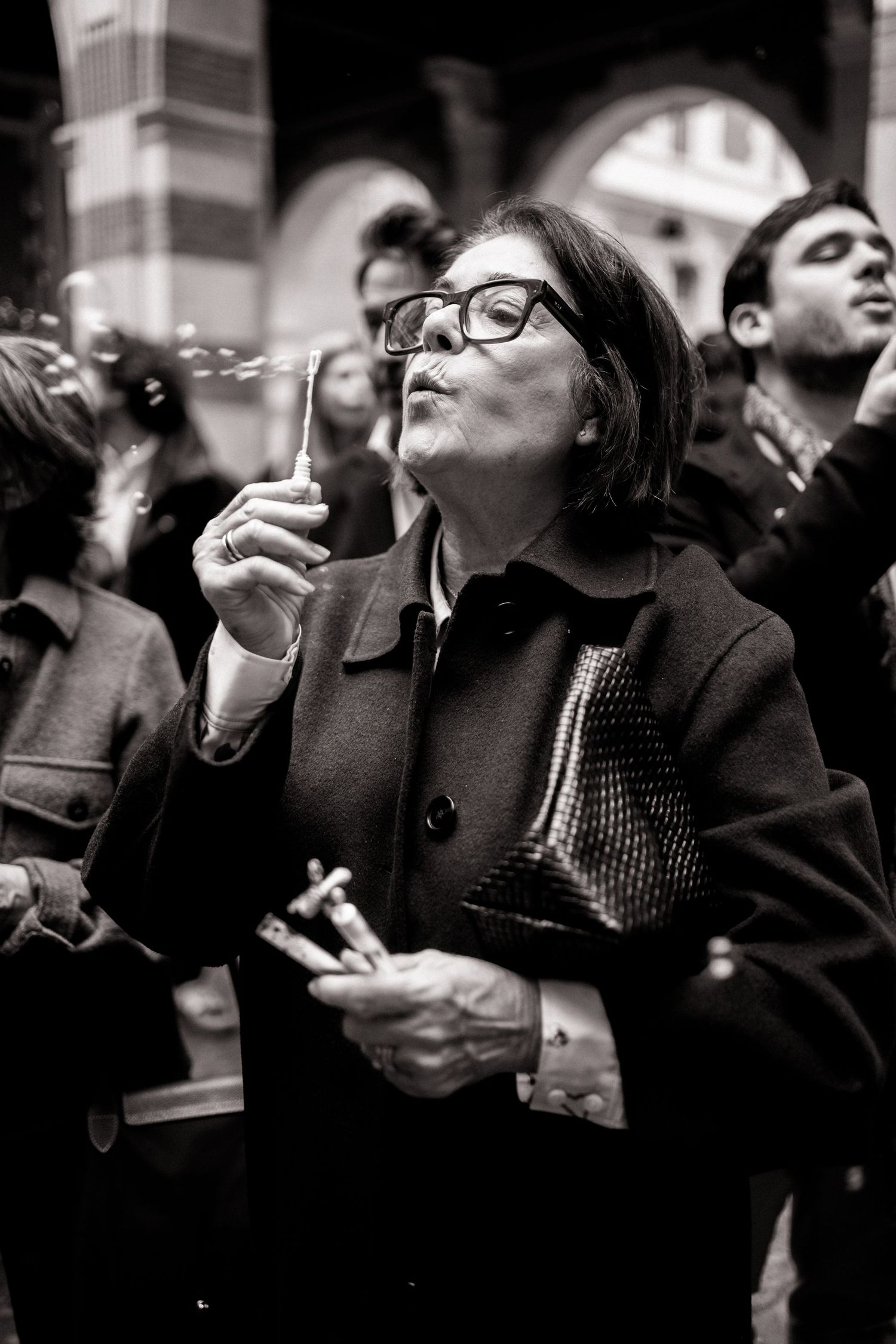 Black and white photo of an older woman blowing bubbles at an outdoor wedding celebration in a crowd.