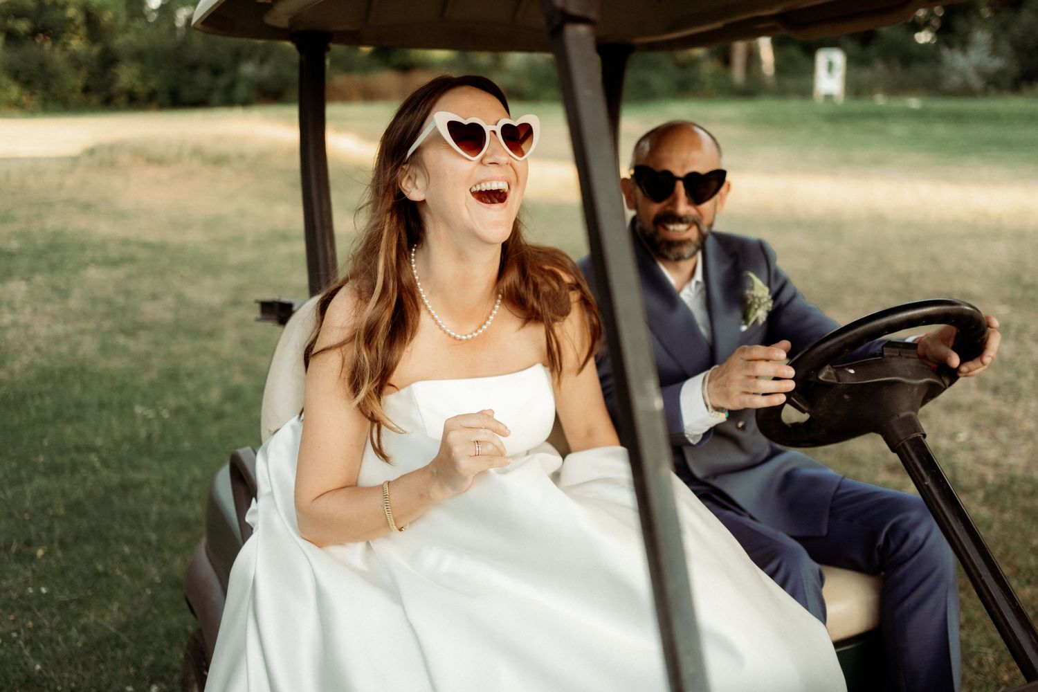 Wedding couple and guests riding in a golf cart through a sunny outdoor venue with joyful expressions.