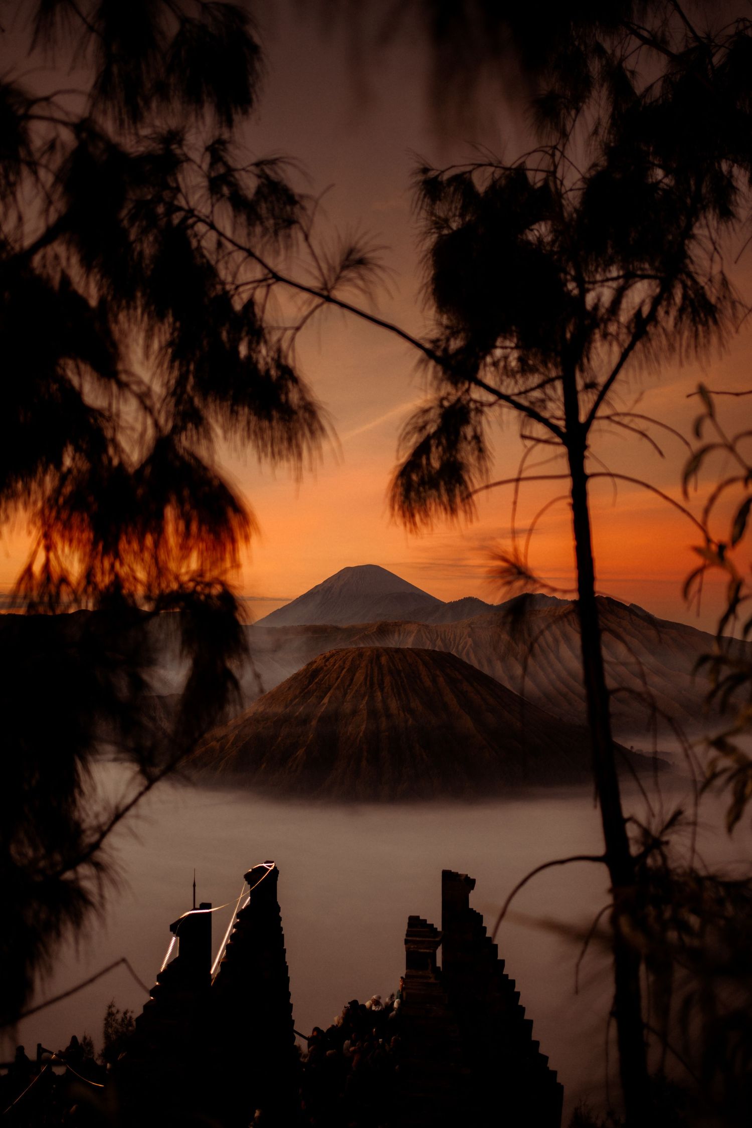 Silhouetted trees frame a dramatic sunrise over Mount Bromo volcano surrounded by misty clouds in Indonesia.