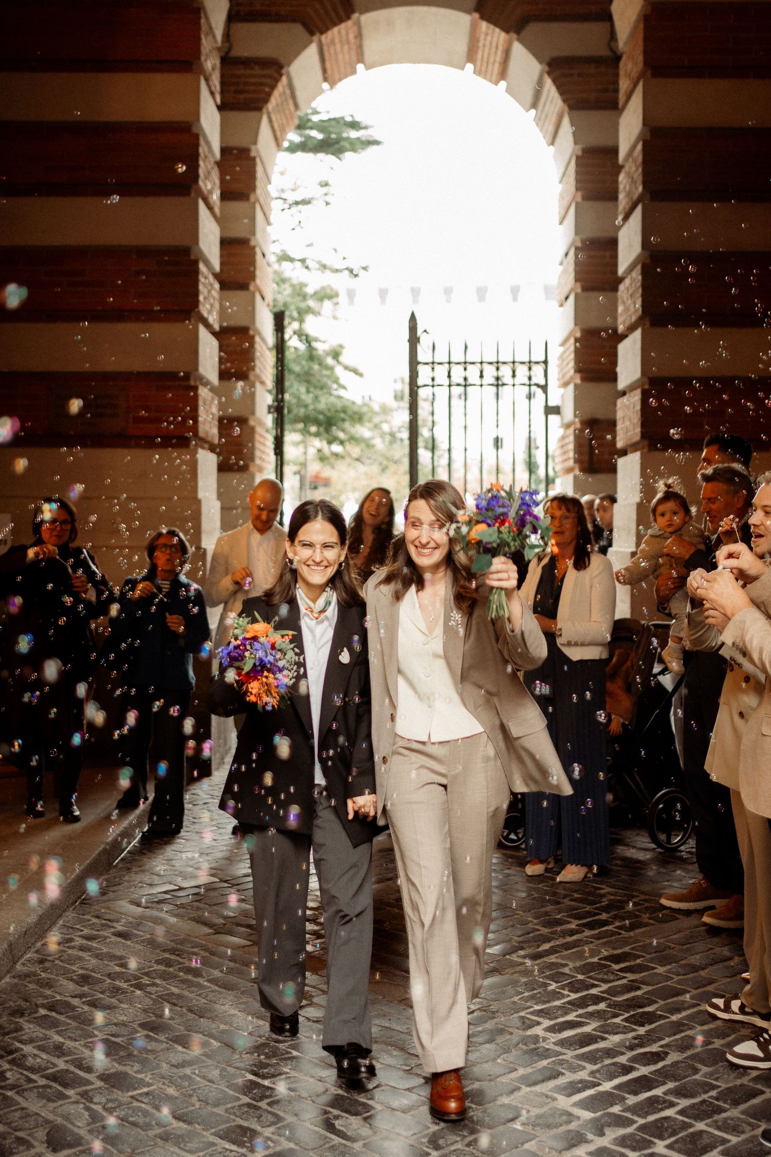 Two brides walking through arch amid confetti shower, guests celebrating their wedding exit.