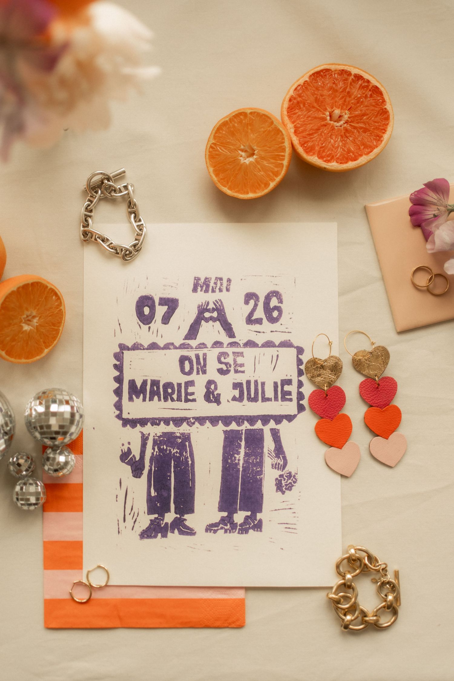 Flat lay of wedding invitation with oranges, jewelry, and heart earrings on neutral background.