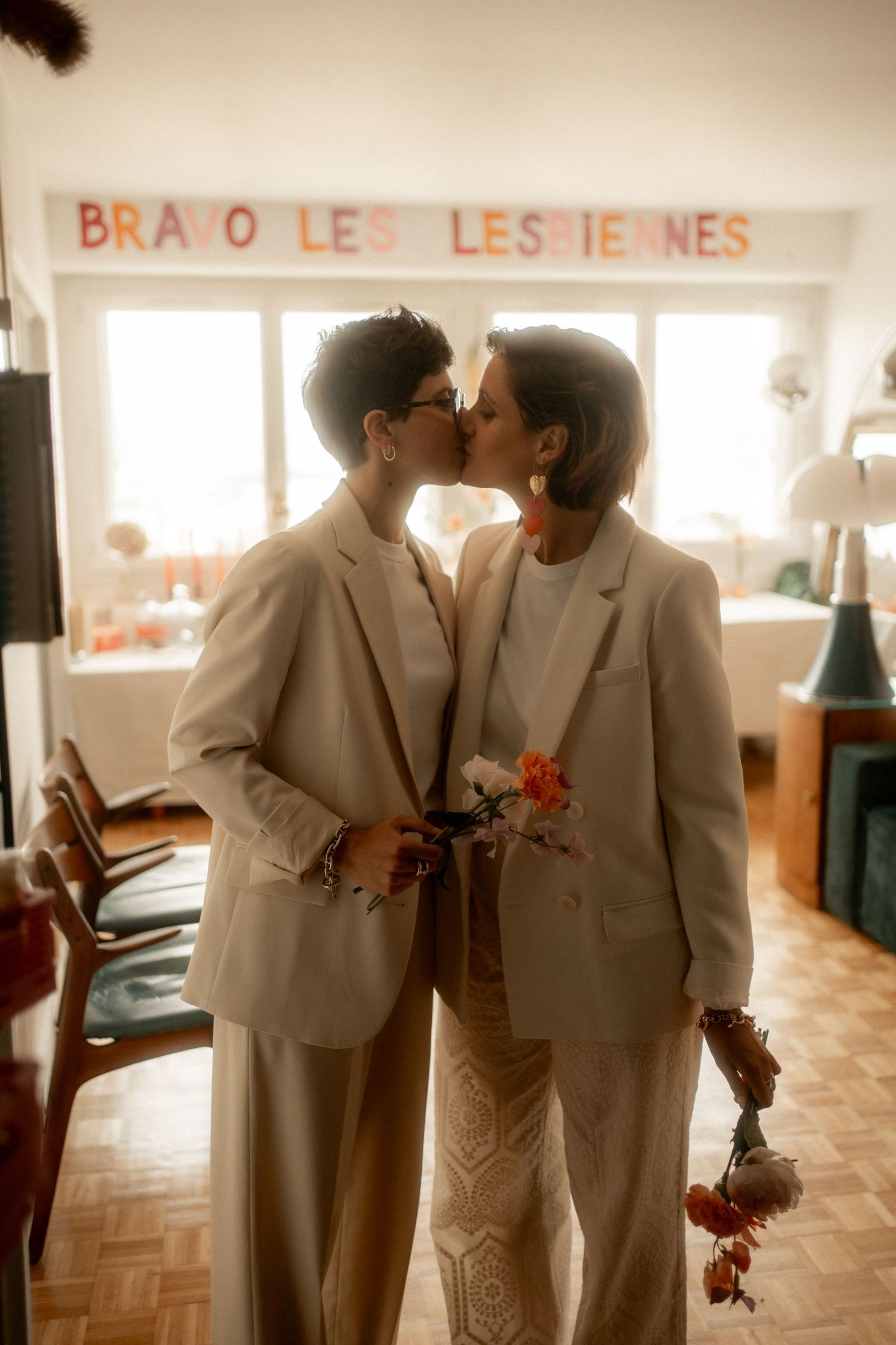 Two women in white suits share a kiss at their wedding, holding flowers under a celebratory banner.