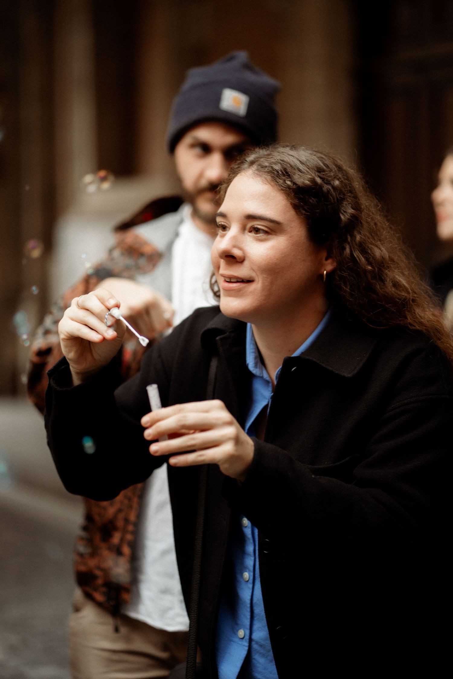 Young woman with curly hair blowing soap bubbles outdoors, wearing black jacket over blue shirt.