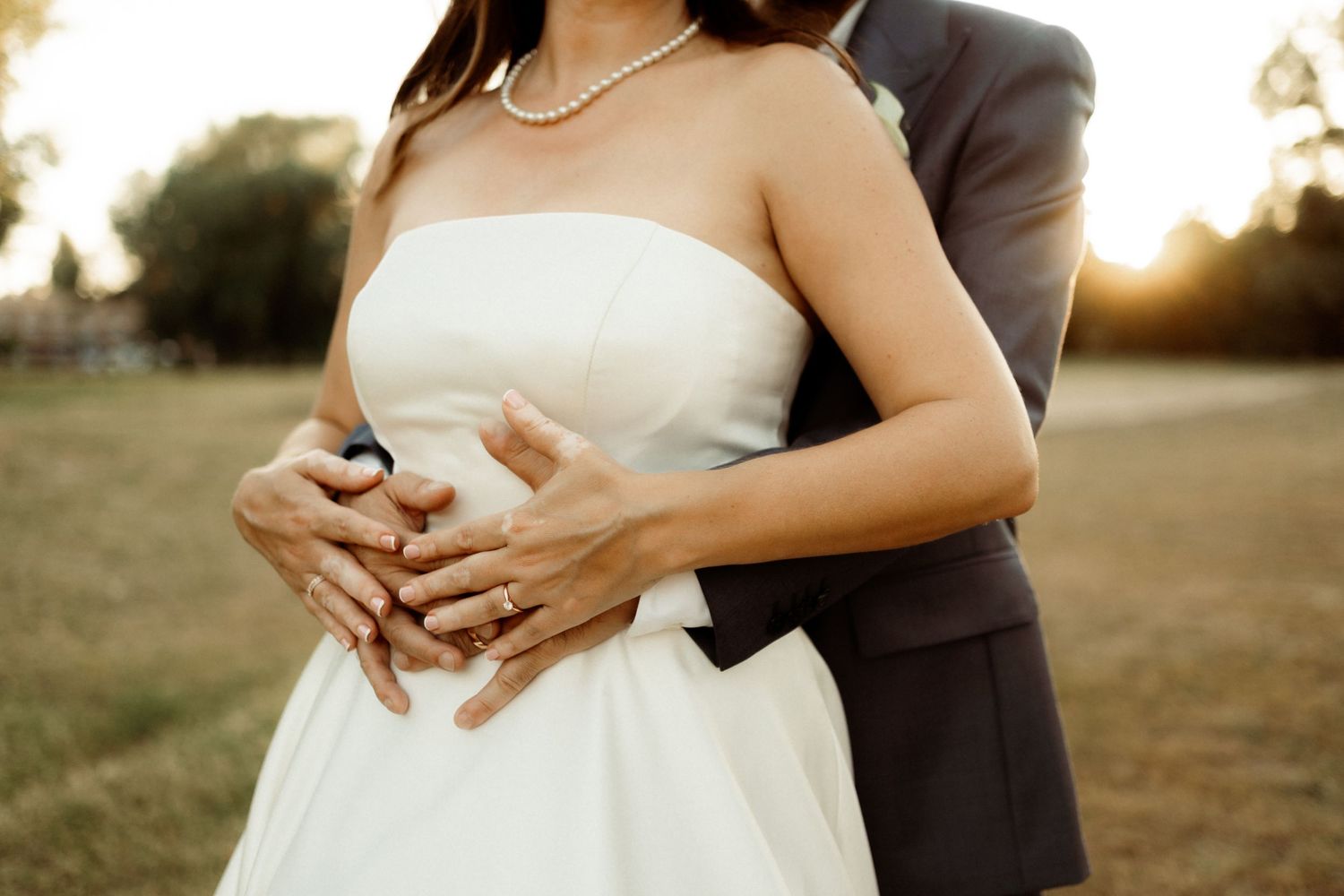 A couple embraces during golden hour sunset, with hands clasped together against a white wedding dress.