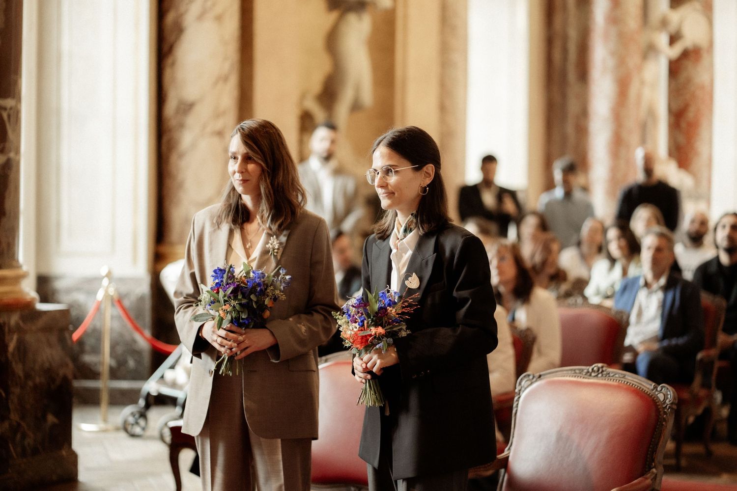 Two women in elegant suits holding colorful flower bouquets stand in an ornate hall during a formal ceremony.