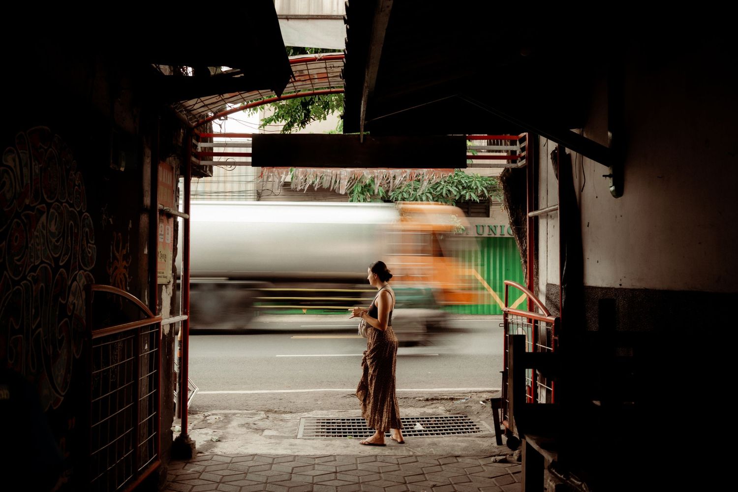 Woman in traditional dress standing in a dark alley watching blurred traffic pass by on a busy street.