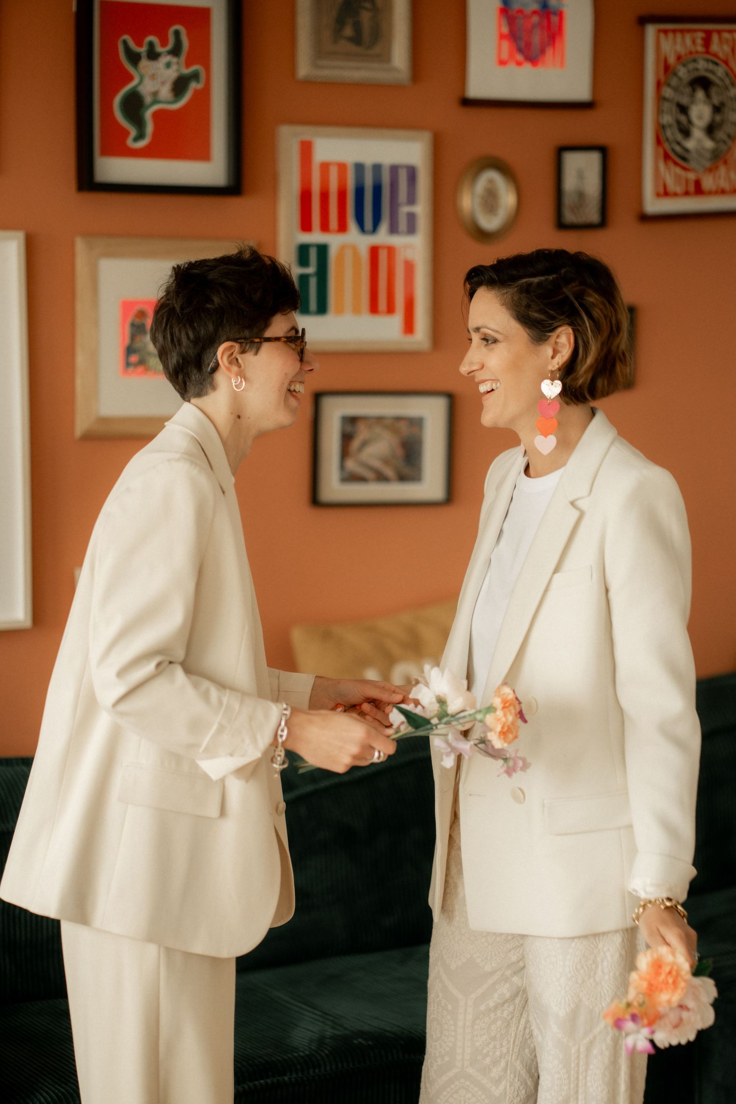 Two brides in white suits hold flowers and smile at each other in front of a colorful gallery wall.