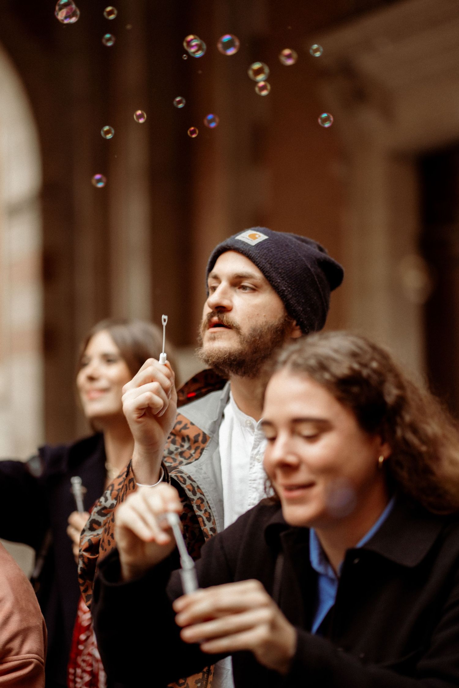 Man in beanie hat blowing soap bubbles outdoors with two women beside him at a historic building.