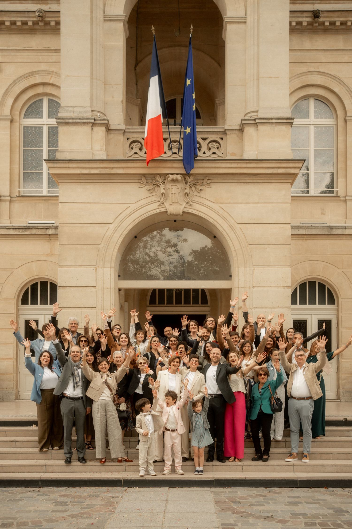 Wedding guests celebrate on steps of a French building with French and EU flags above.