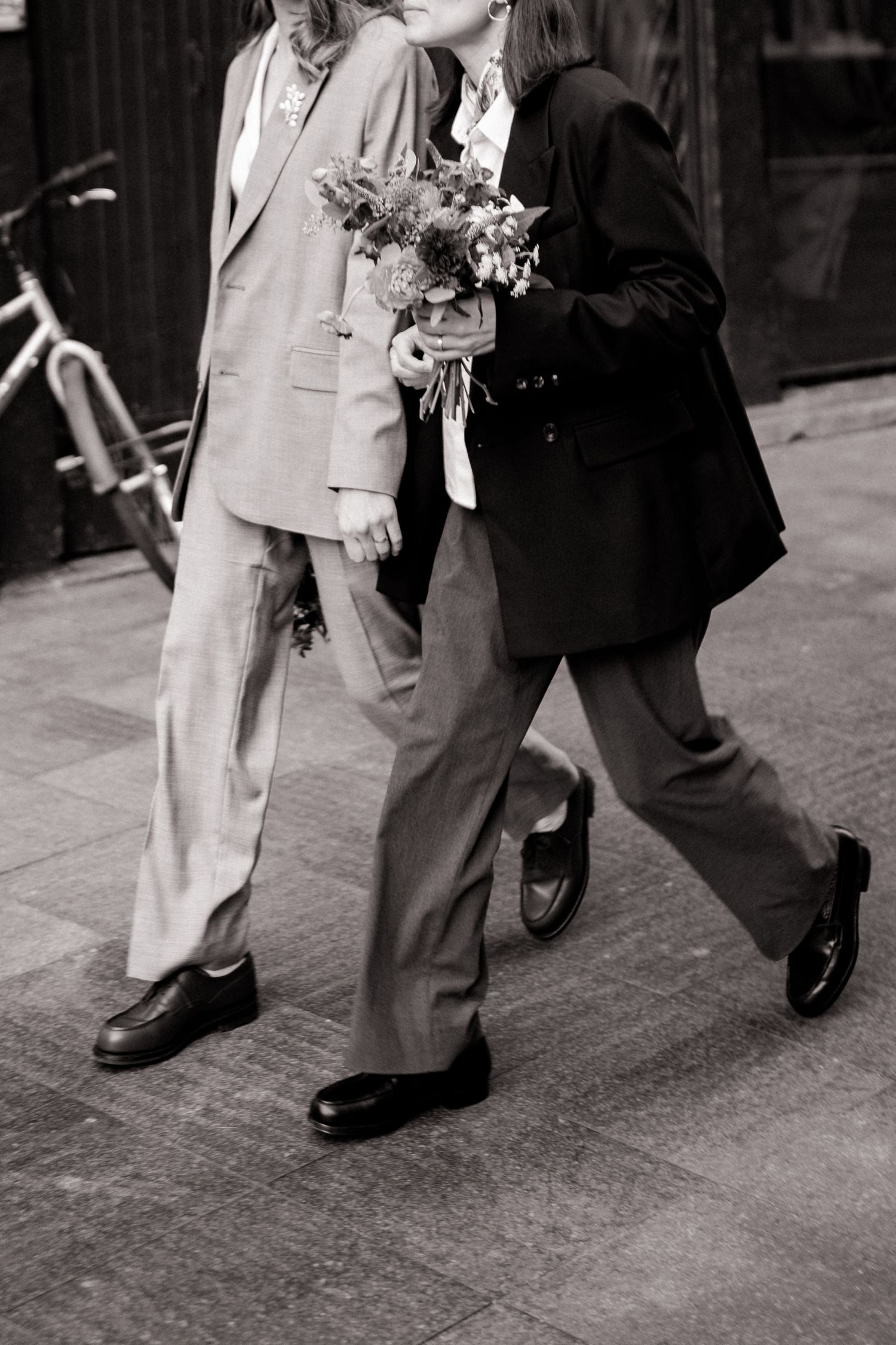 Black and white photo of two people in suits walking together, one holding a wedding bouquet.