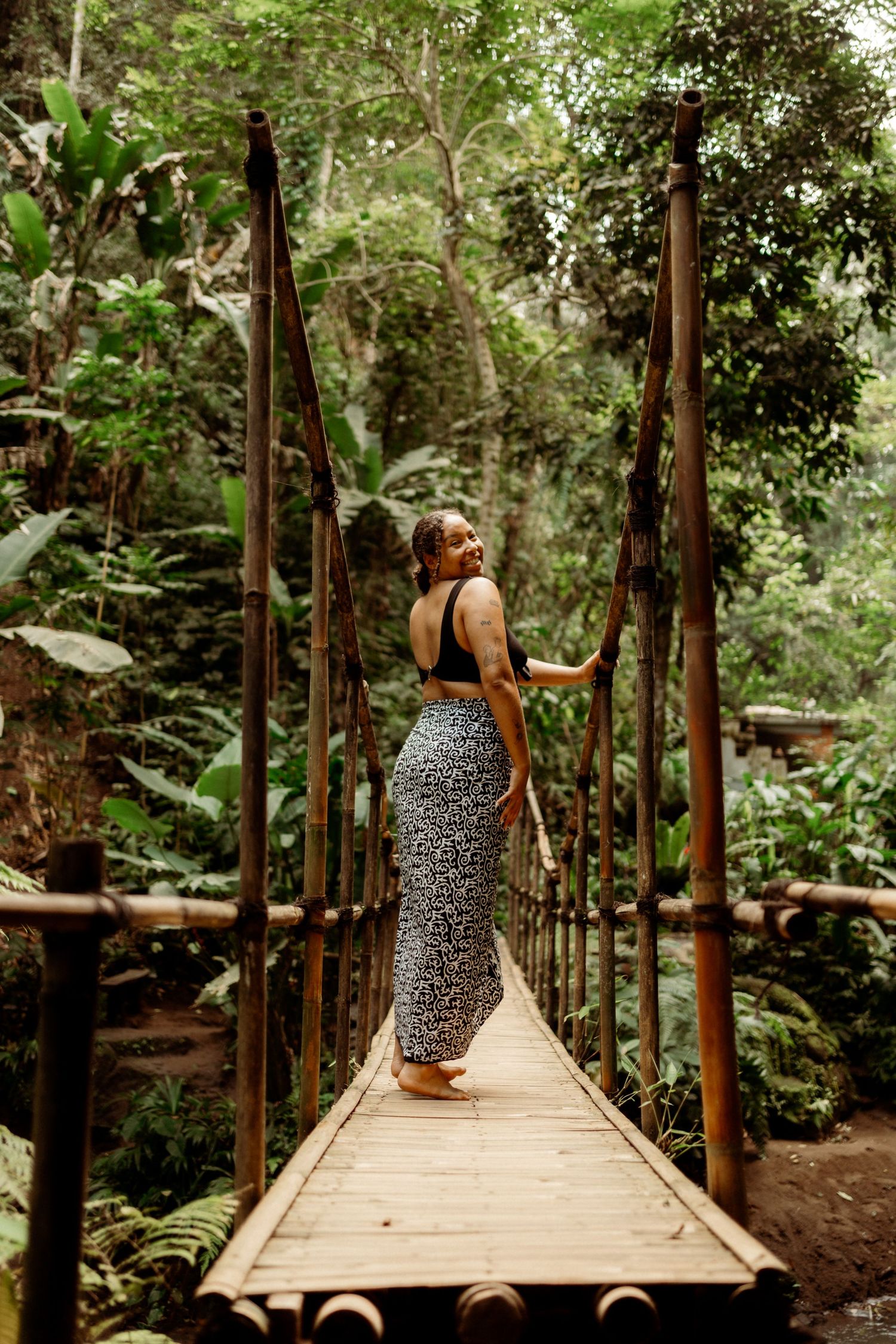 Woman in floral skirt stands on wooden suspension bridge surrounded by lush tropical jungle greenery.