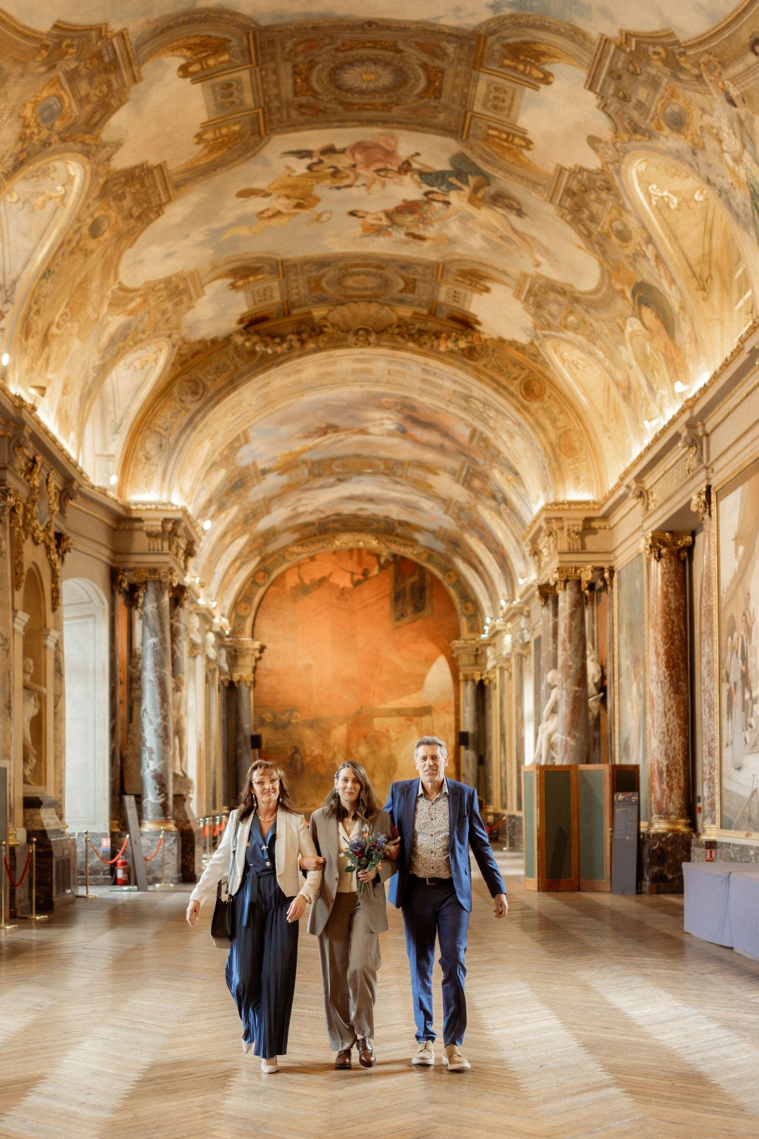 Three people walking through an ornate baroque gallery hall with elaborate frescoed ceiling and classical architecture.