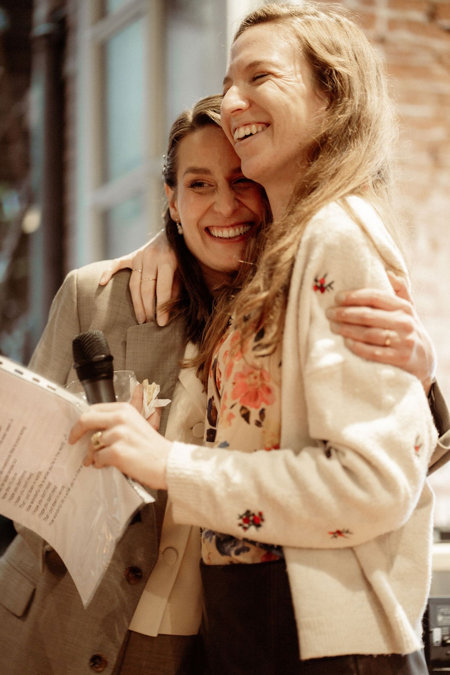 Two women laughing and hugging warmly, one holding a microphone and papers at an indoor event.