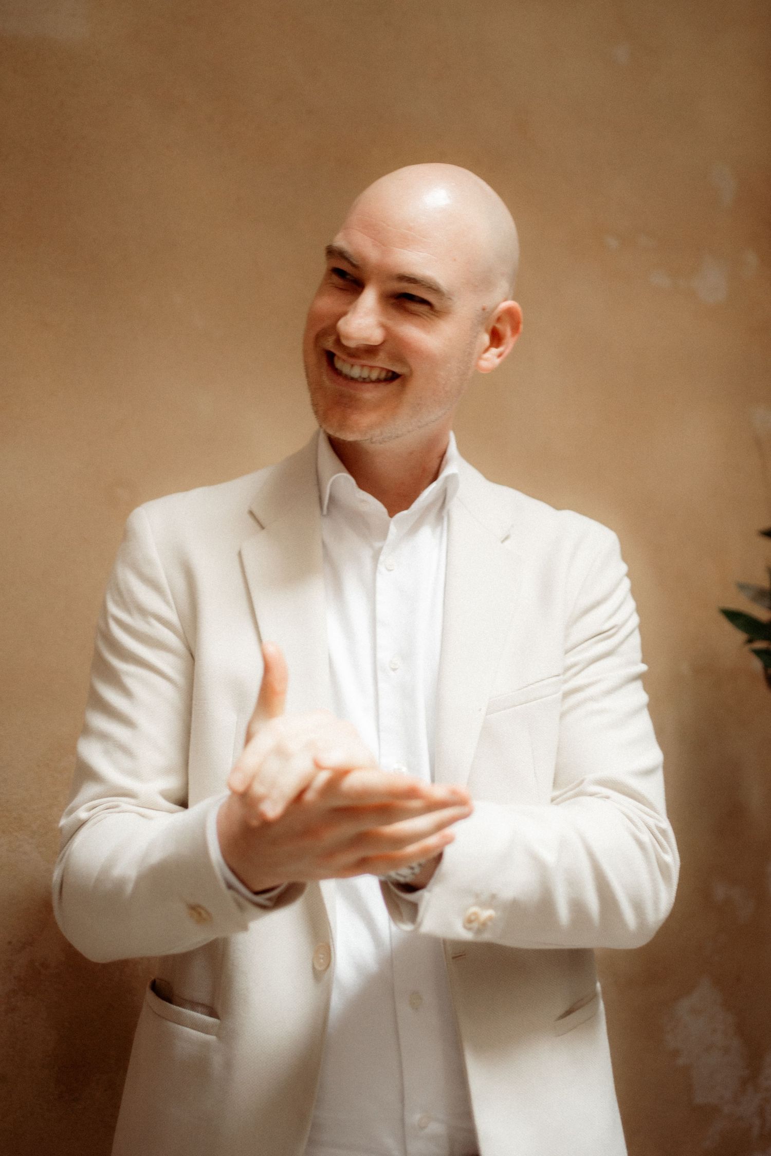 Smiling bald man in white blazer clapping hands against beige wall background.