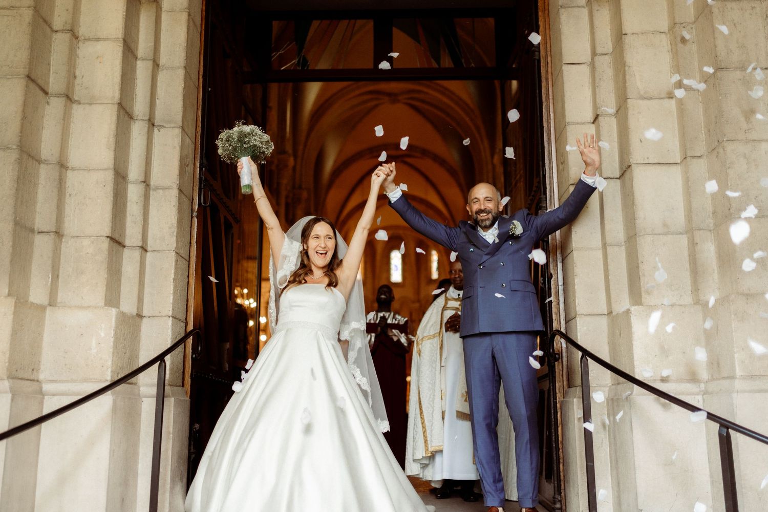 Newlyweds celebrate joyfully with raised arms on church steps while confetti falls around them.