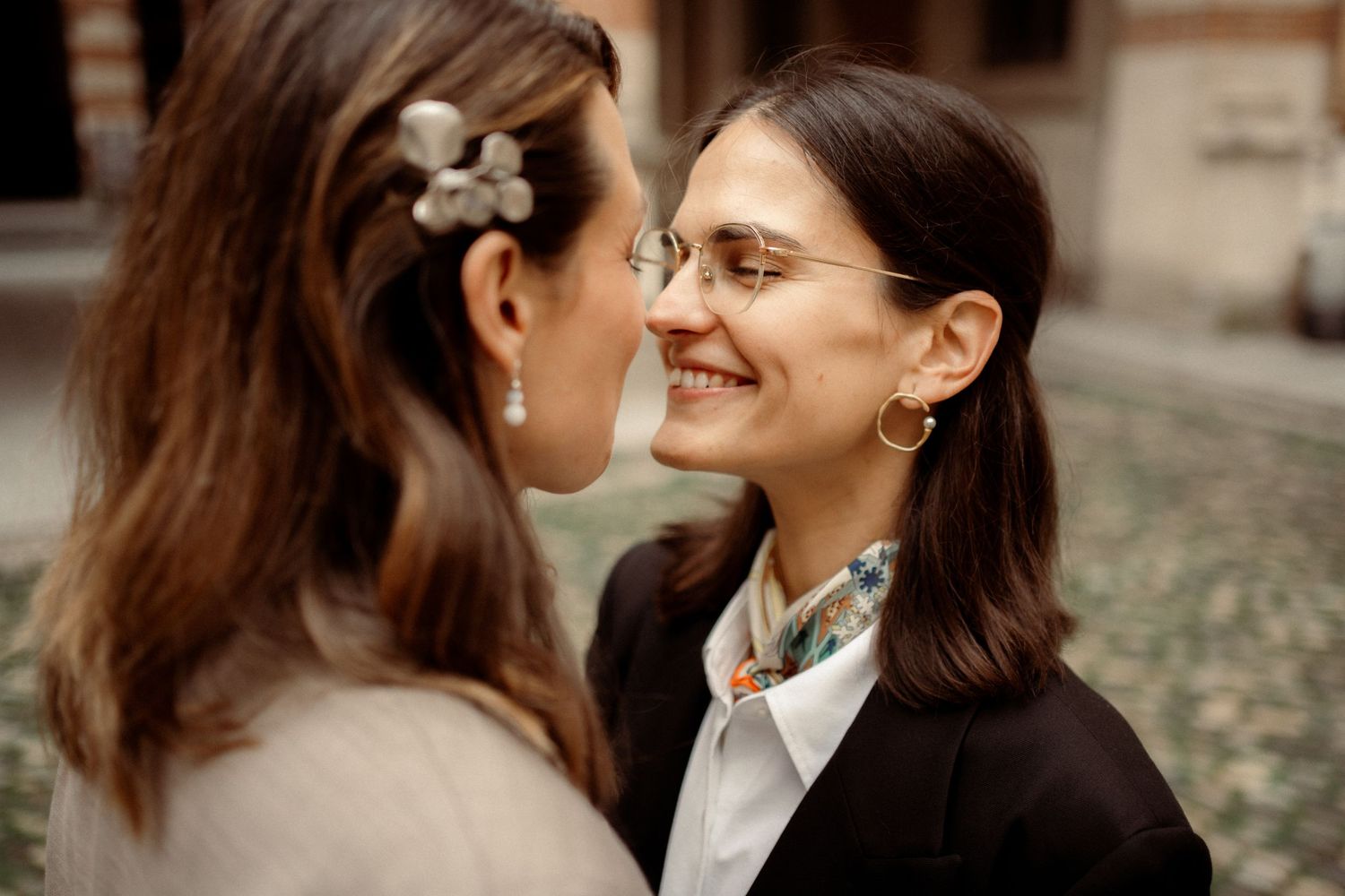 Two women smiling and facing each other closely, one wearing glasses and a black blazer with a decorative scarf.