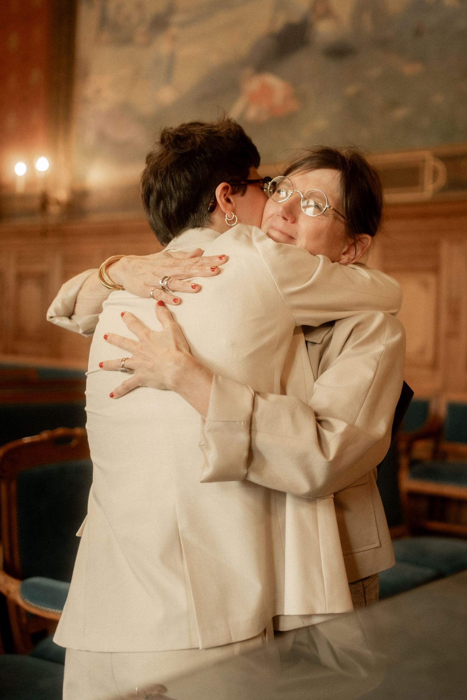 Two women in white suits share an emotional hug during a wedding ceremony indoors.