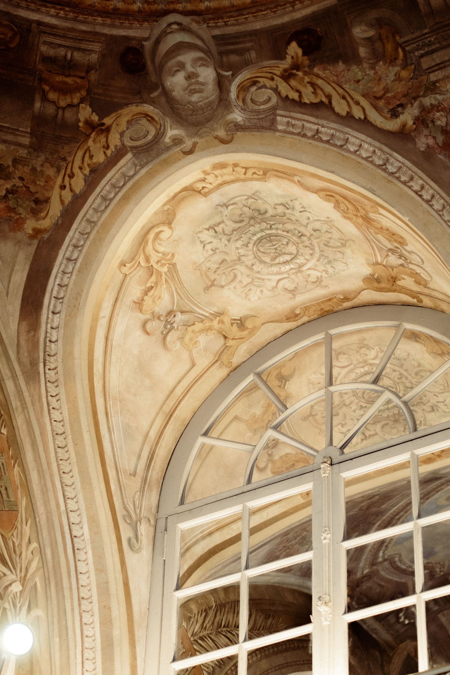 Ornate Baroque architectural detail featuring gilded stucco moldings, floral relief, and arched window with sculpted face above.