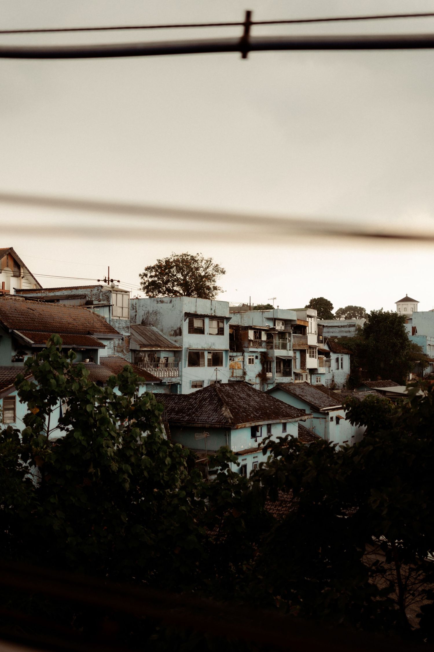 Rows of aged white buildings with tiled rooftops viewed through wire fence at dusk.