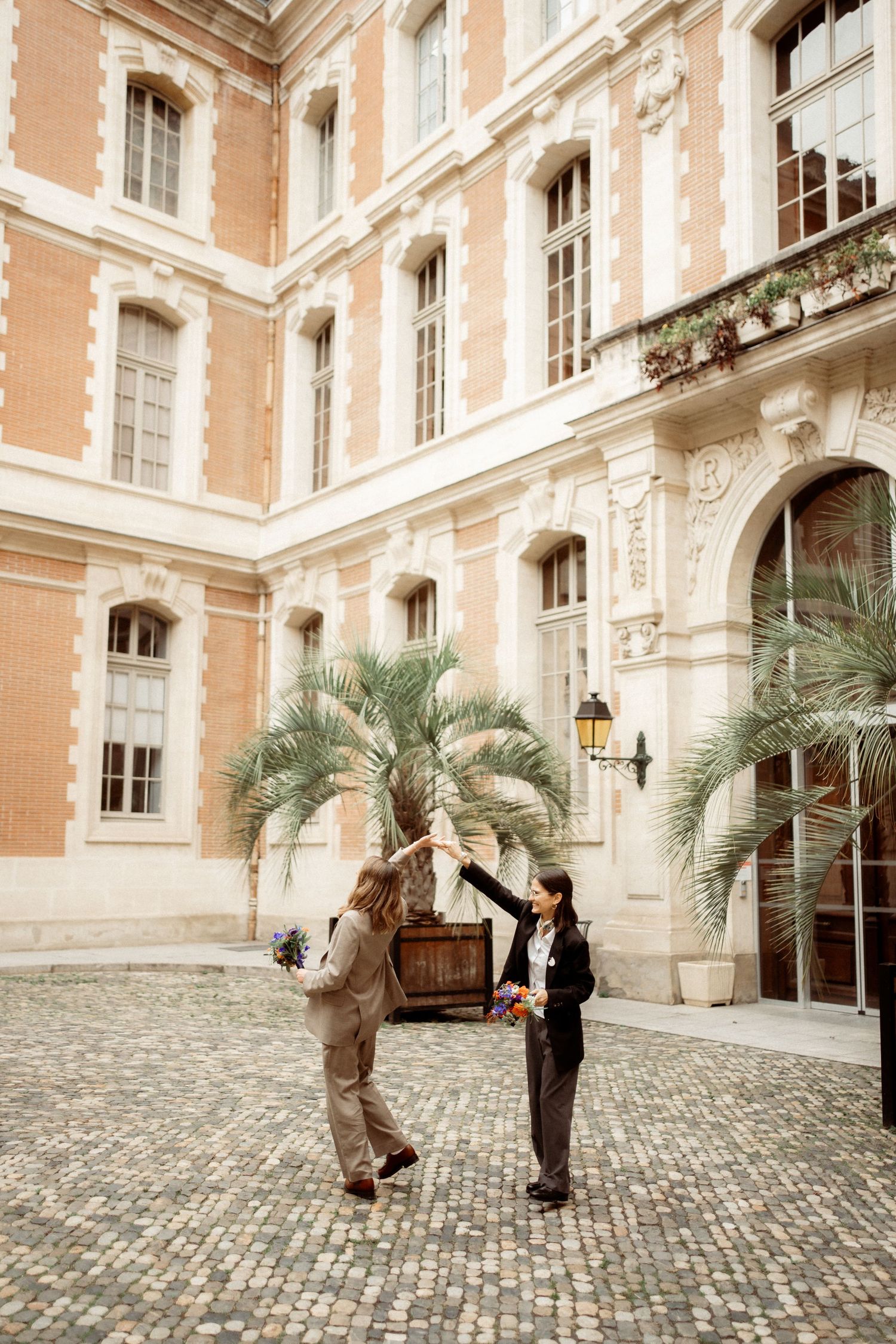 Two people high-fiving in elegant cobblestone courtyard of classic French building with palm trees.