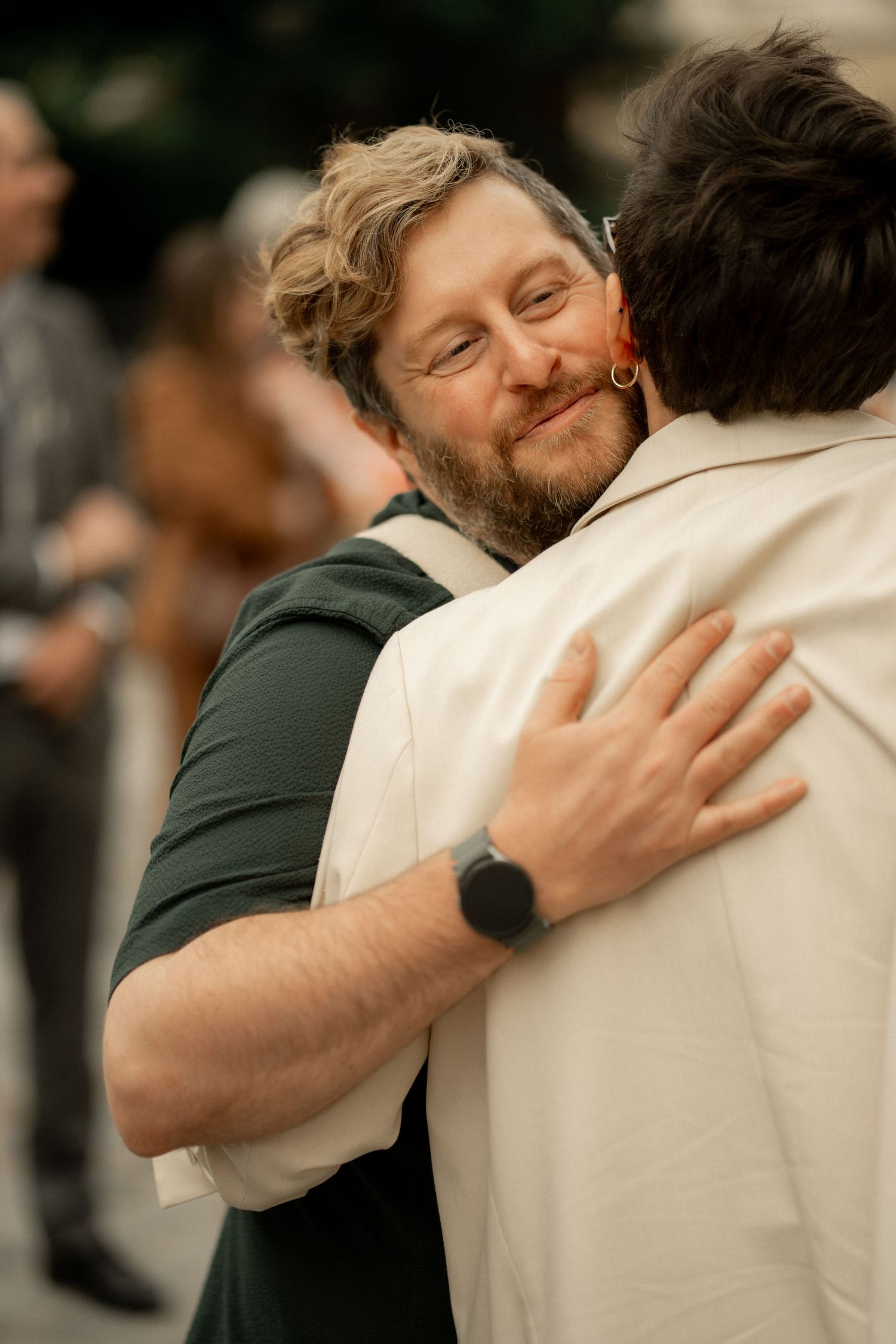 Bearded man with curly hair smiling warmly while embracing someone at an outdoor social gathering.