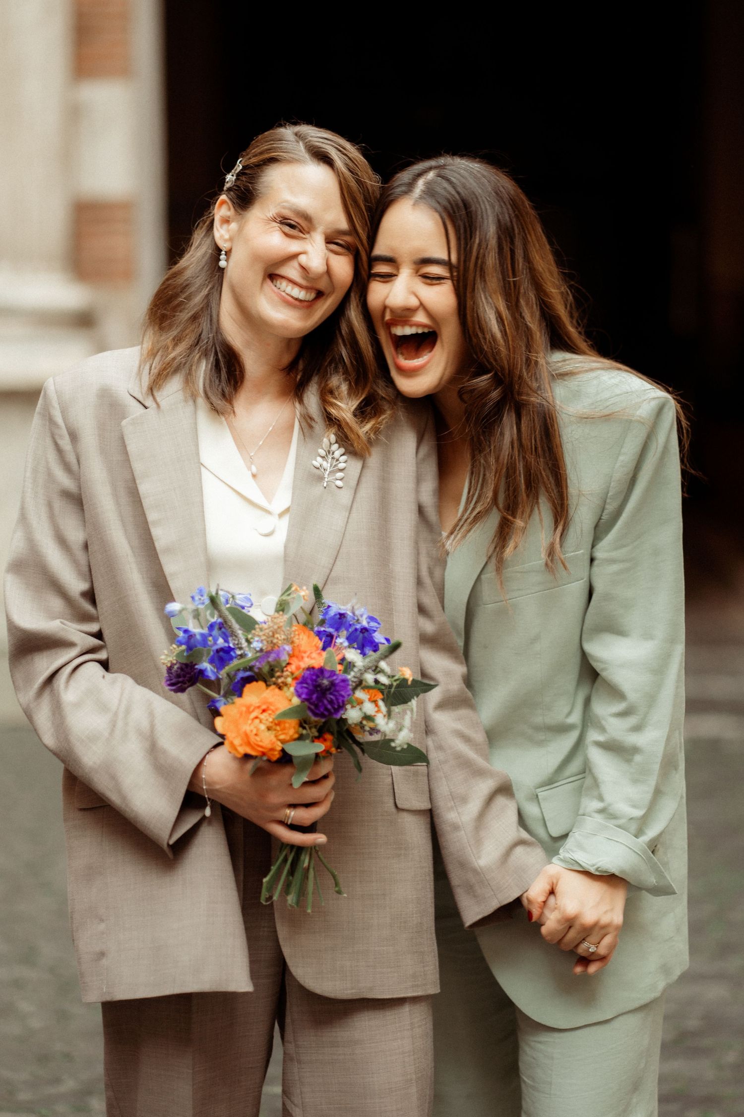 Two brides in suits laughing joyfully, one holding a colorful bouquet of orange and purple flowers.