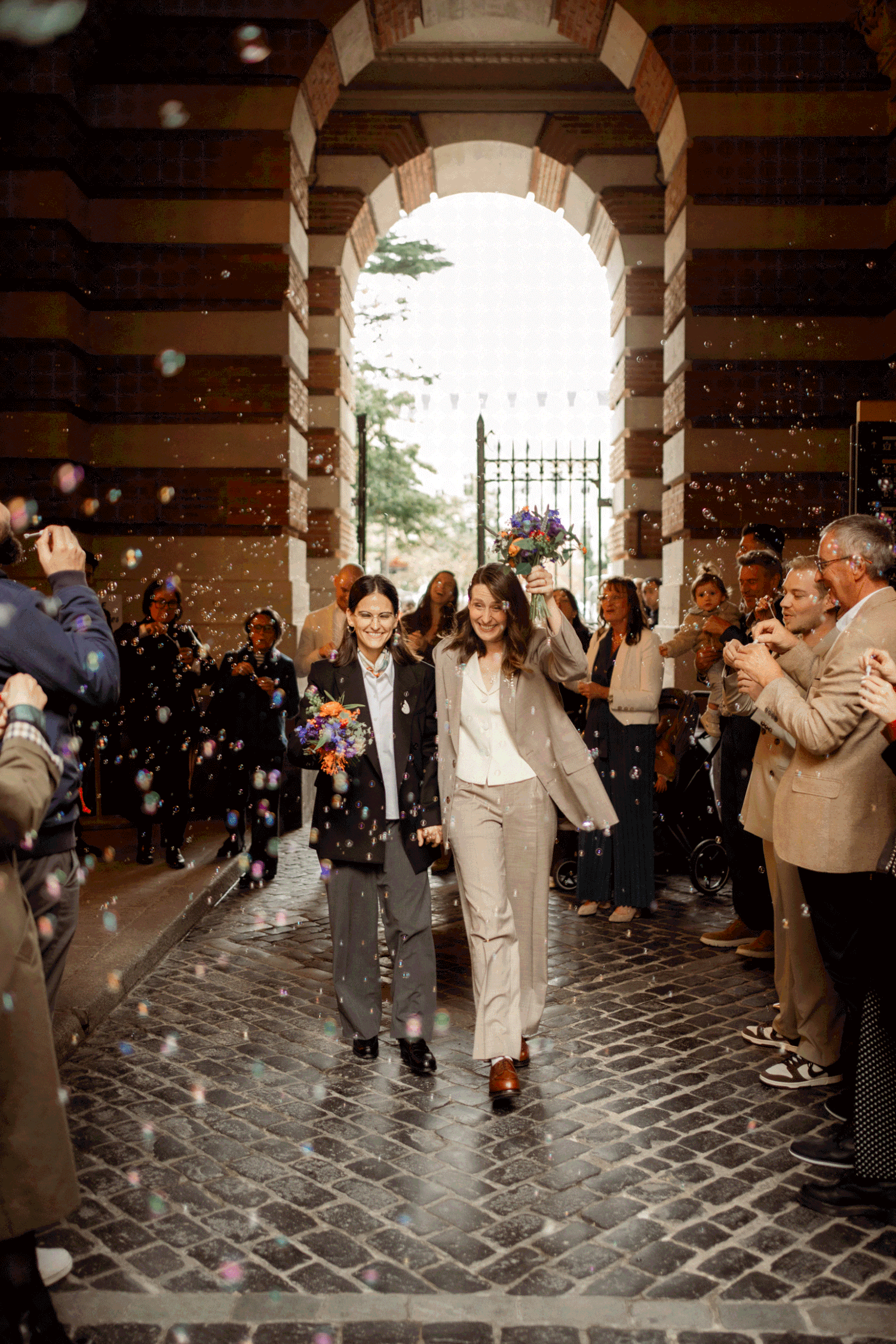 Two brides walk through confetti shower under stone archway, guests celebrating their wedding exit.