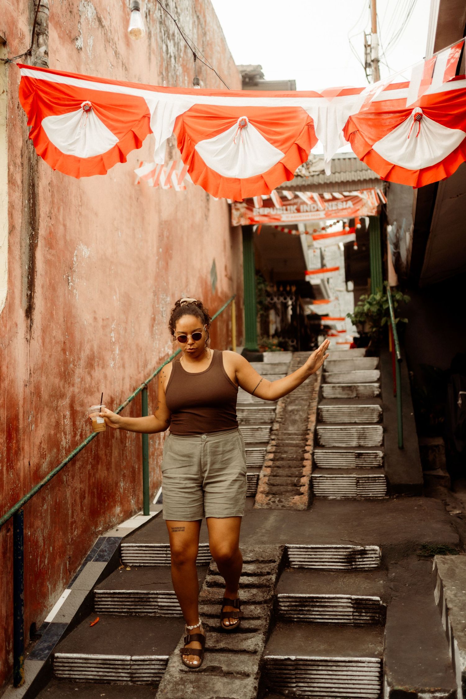 Woman in brown tank top and shorts stands on steps in colorful alley with red and white bunting.