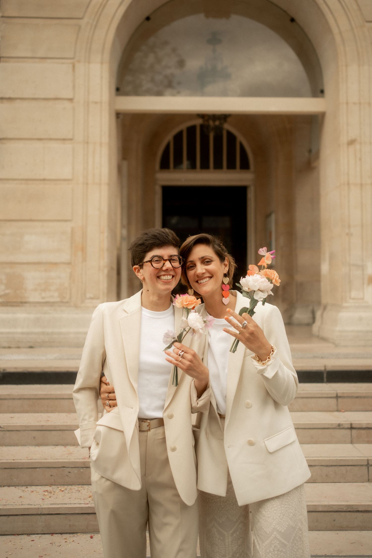 Two women in elegant cream suits holding flowers and smiling on steps of a grand building.