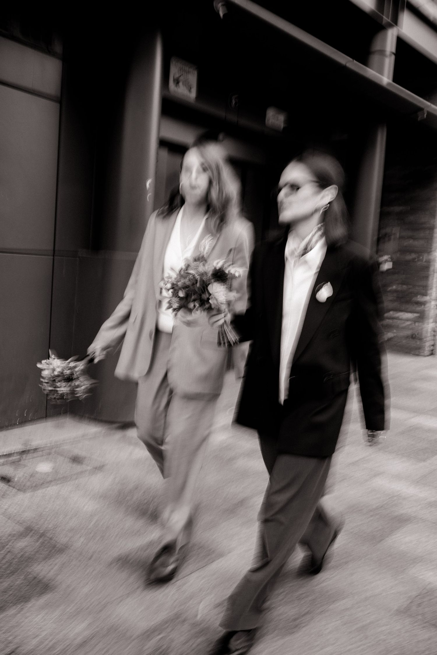 Blurred black and white photo of two brides in suits walking together holding wedding bouquets.