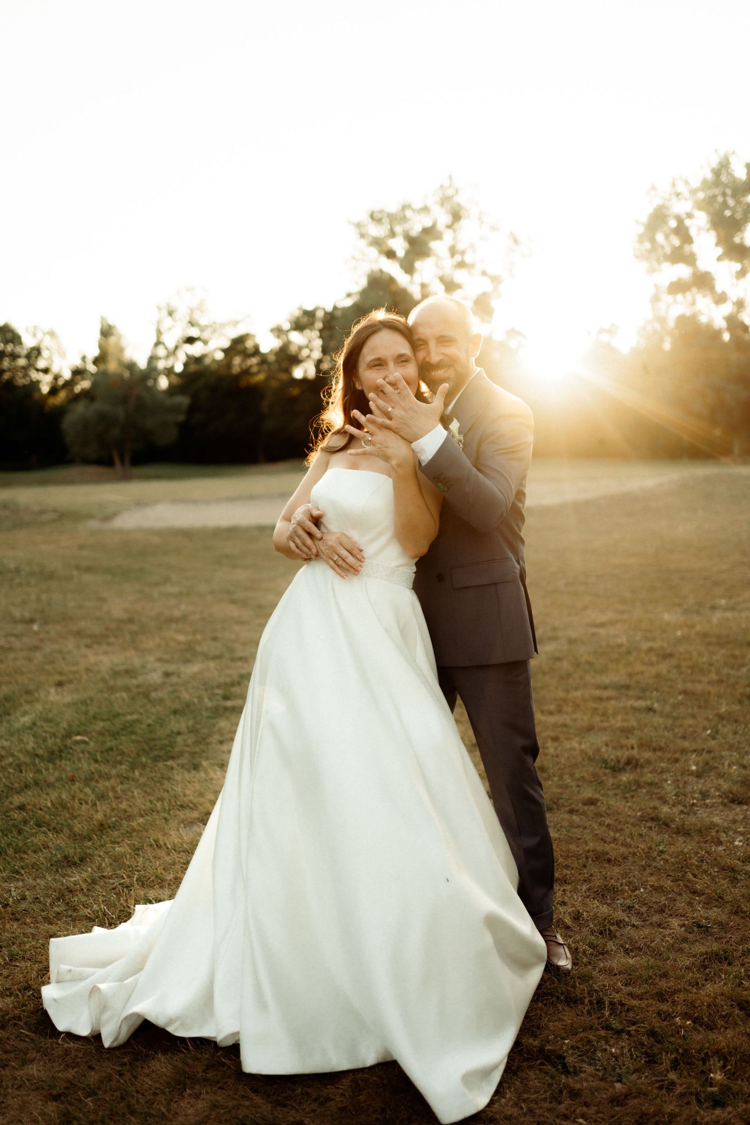 A couple embraces in a romantic sunset moment on their wedding day, with golden light illuminating the outdoor setting.