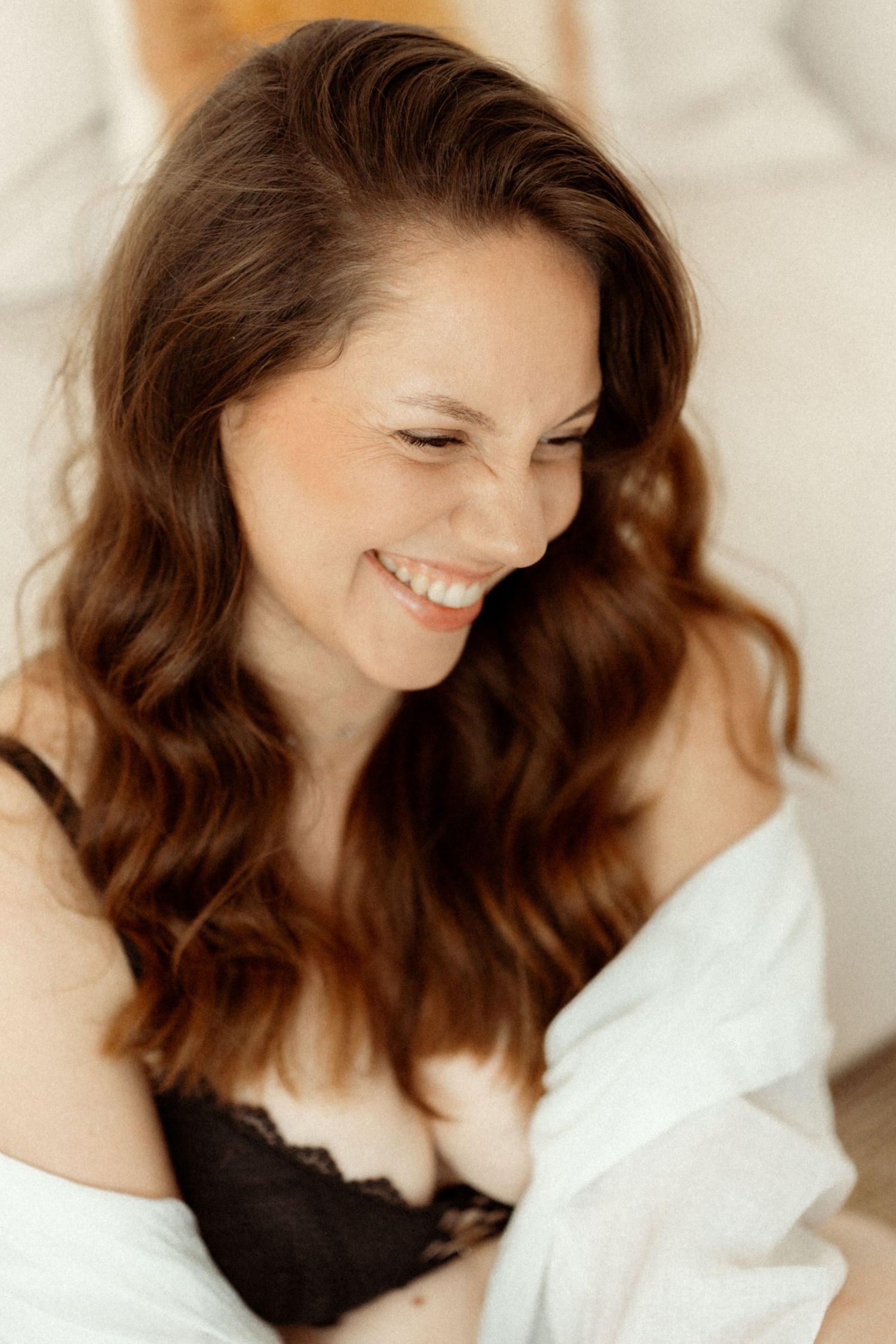 Woman with wavy brown hair and natural makeup smiling in soft lighting against white background.