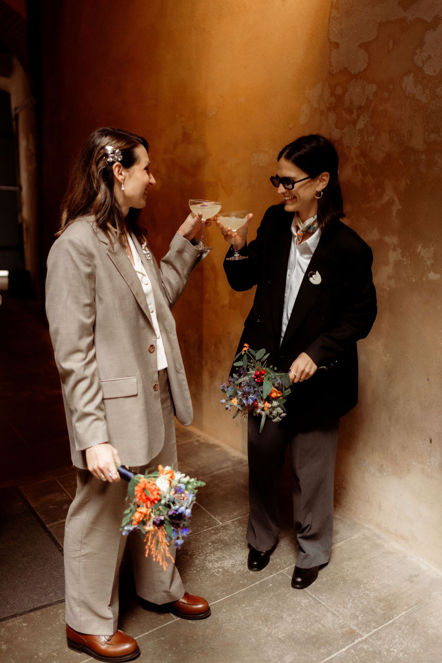 Two brides in elegant suits toasting with cocktail glasses while holding colorful wedding bouquets indoors.