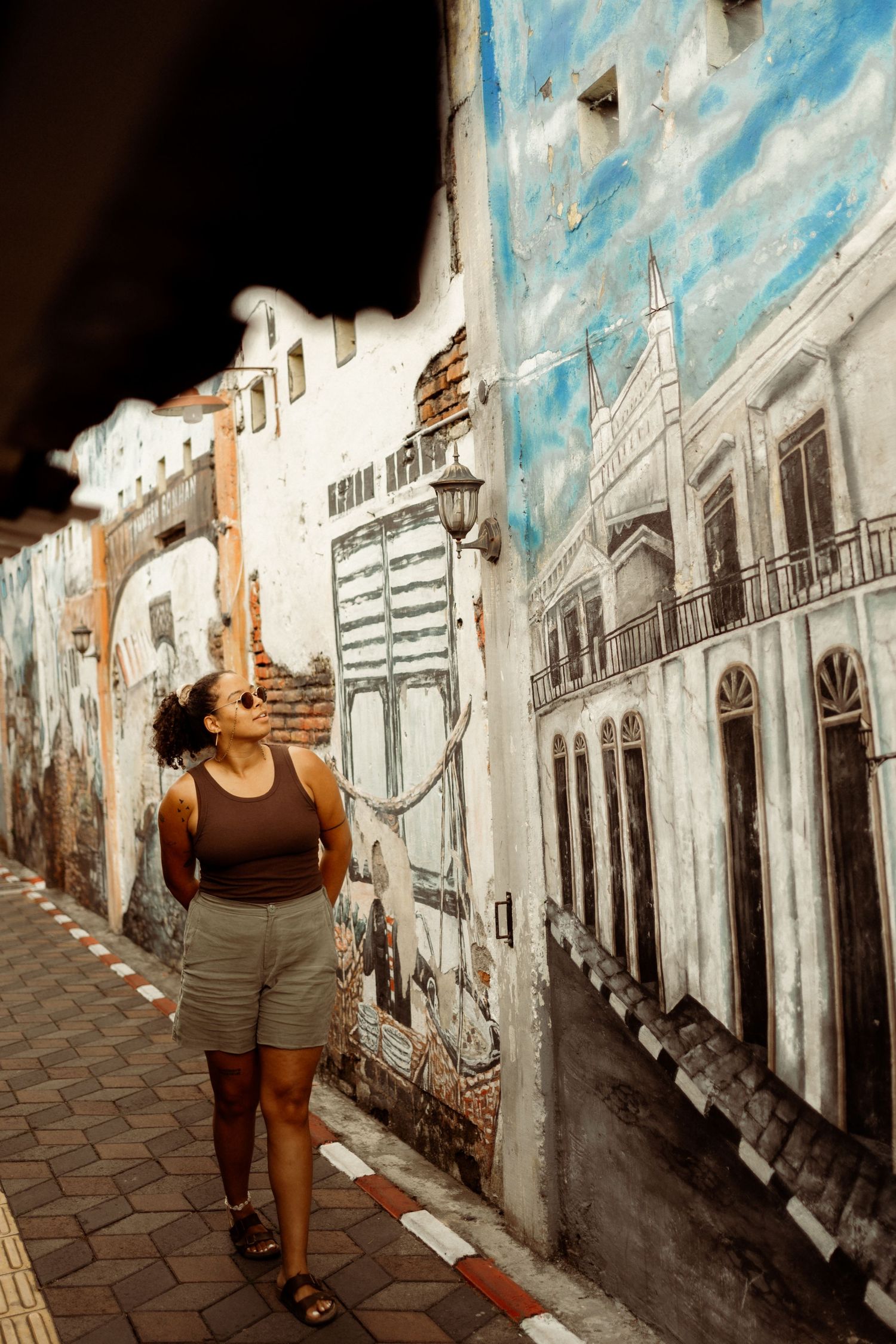 Woman in sunglasses admiring detailed street art mural of historic buildings on an urban wall.