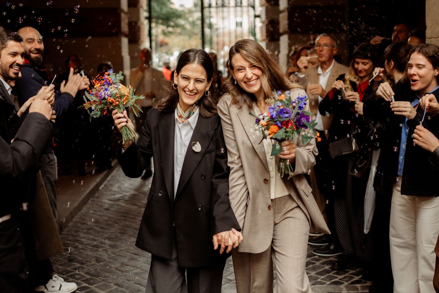Two brides in suits hold hands and bouquets while guests celebrate with confetti on a cobblestone street.