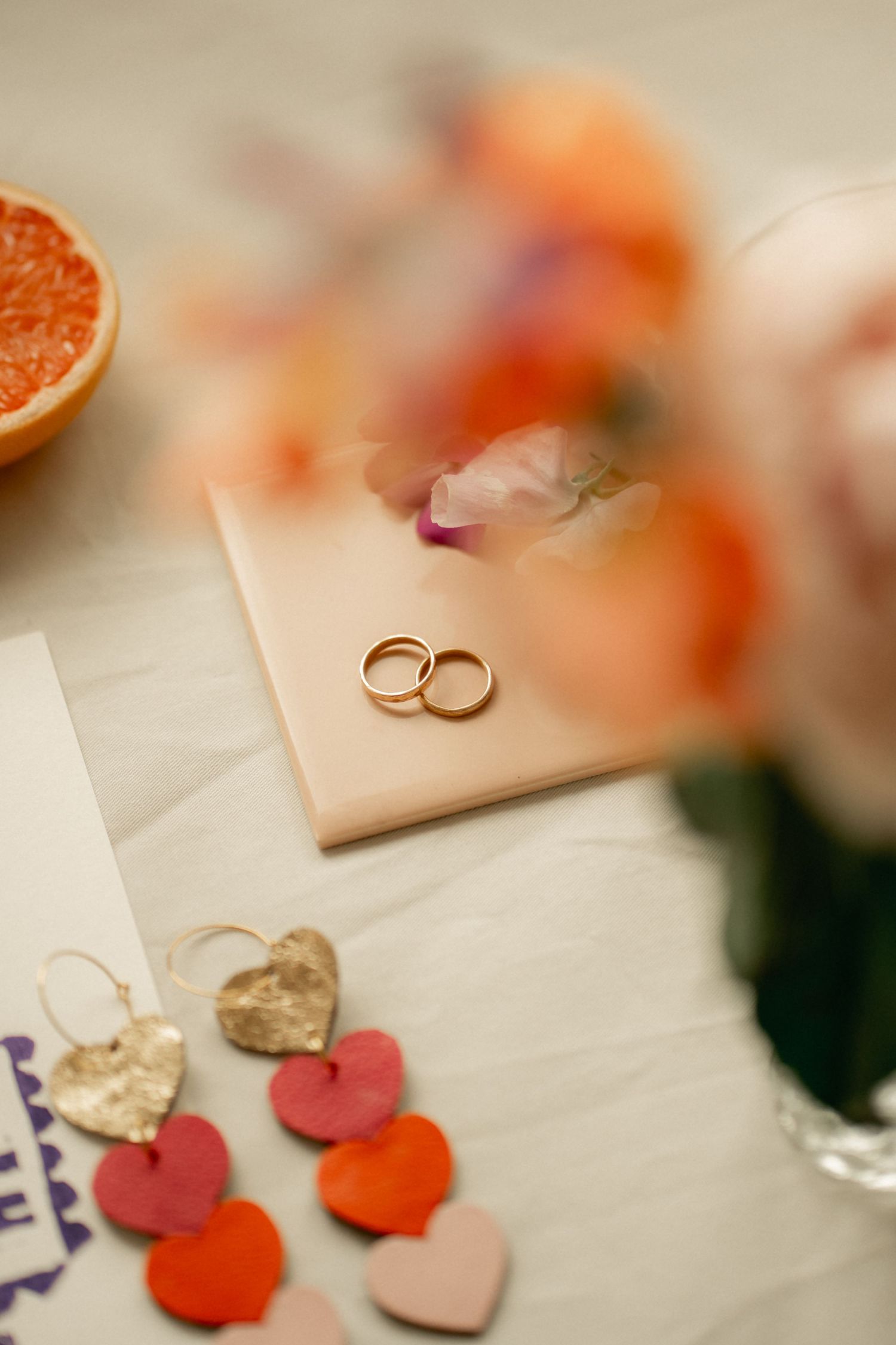 Gold wedding rings resting on a pink tile surrounded by heart-shaped earrings, flowers, and a halved grapefruit on a white surface.