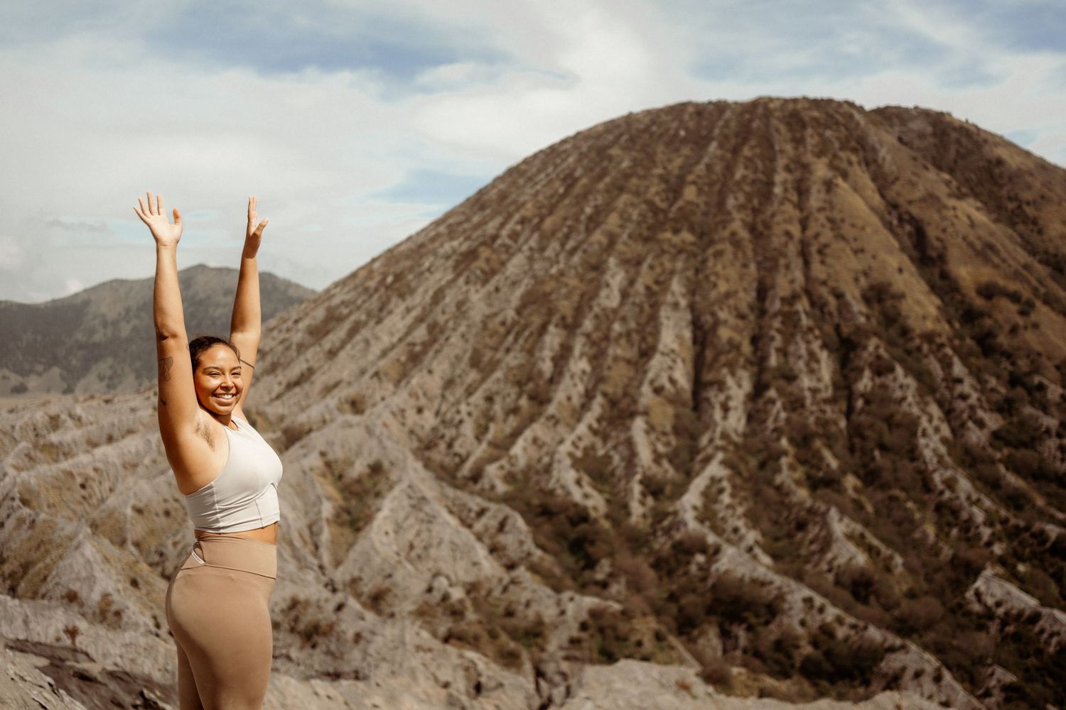 Smiling woman with arms raised stands before Mount Batok volcanic crater in East Java Indonesia.