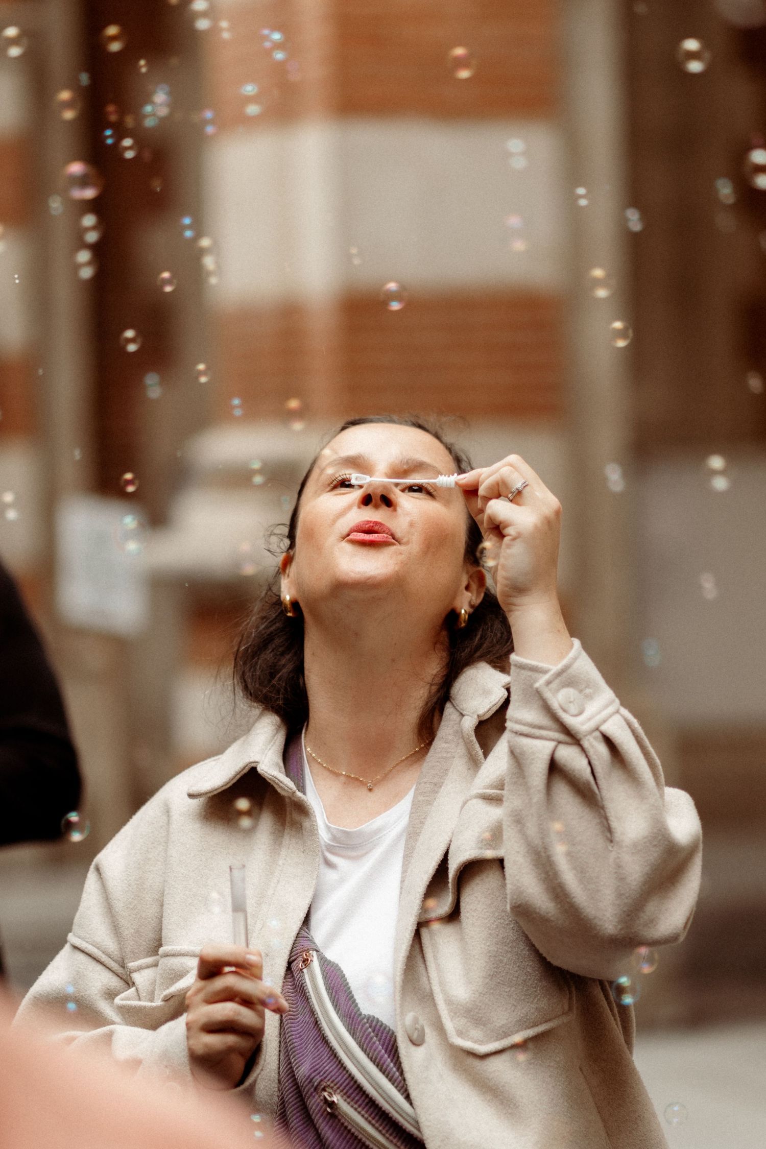Young woman blowing soap bubbles outdoors, wearing a beige jacket and crossbody bag, surrounded by floating bubbles.